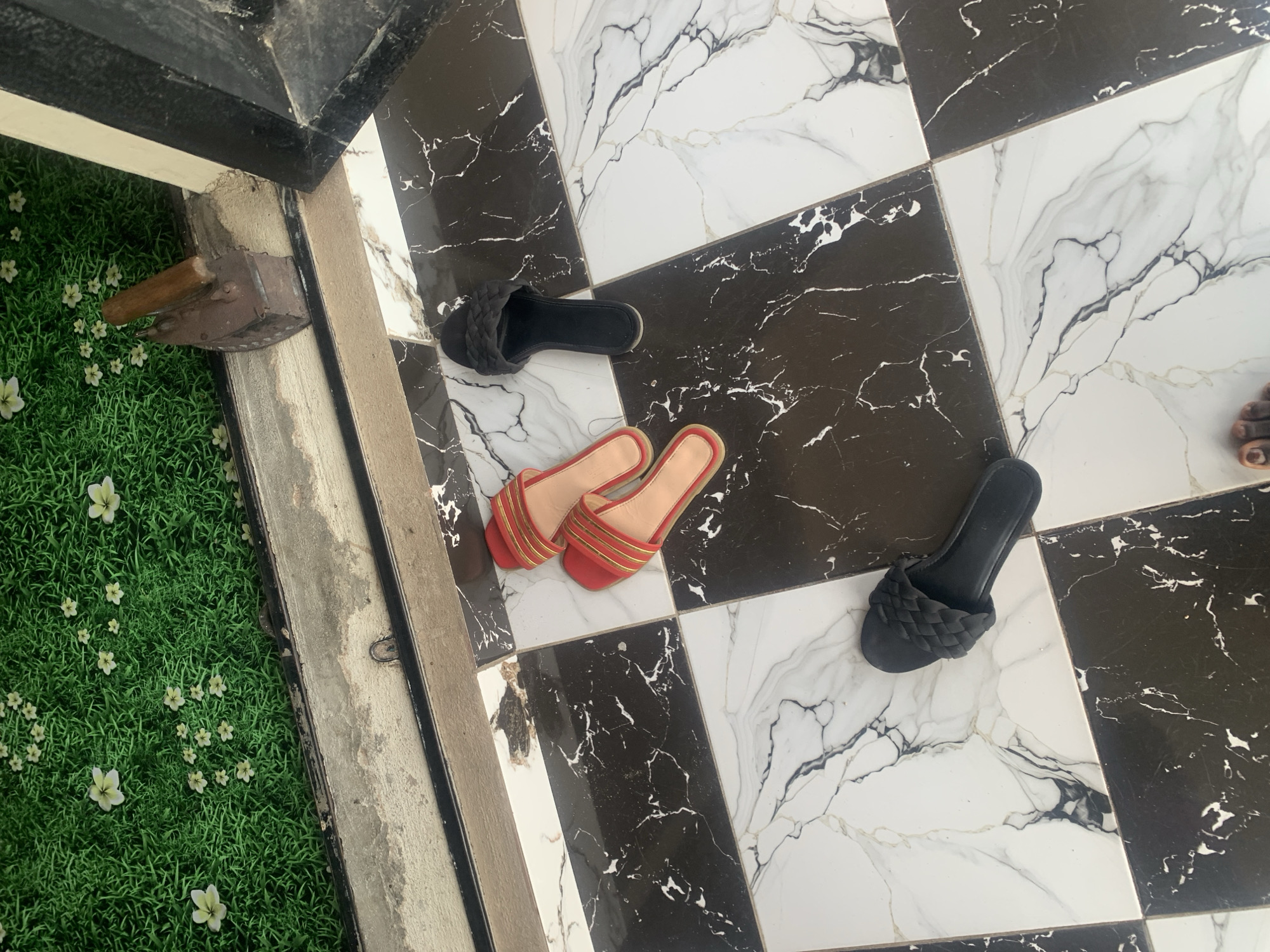 In Jauro Yinu, Nigeria, a pair of red flat sandals with gold accents is placed on a black and white marble tiled floor, alongside a black braided slide sandal and another black slide sandal. The scene is located at an entrance, with a dusty, weathered concrete threshold separating the tiled floor from a patch of artificial green grass with scattered white flowers. An old, rusty metal iron with a wooden handle rests on the threshold. A person's left foot, wearing a black slide sandal, is visible in the lower left corner of the image, suggesting recent presence.
