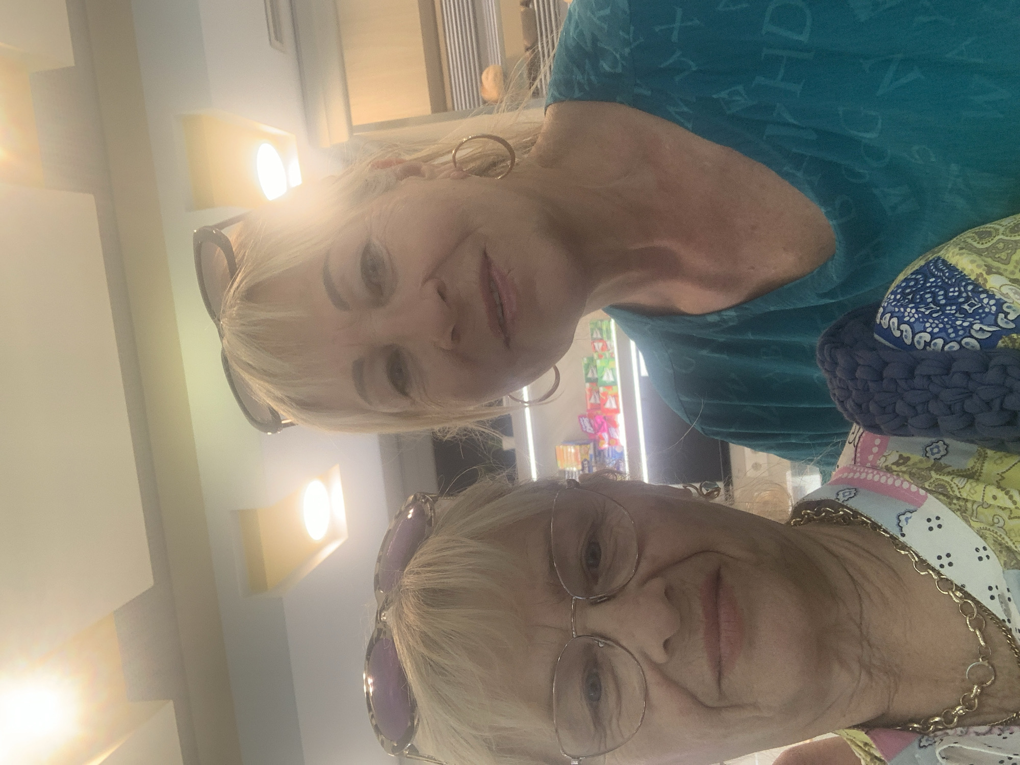 Two women pose for a selfie in what appears to be a commercial establishment with shelves stocked with products. The woman on the left wears glasses, a patterned shirt, and a gold chain. The woman on the right has blonde hair and wears a teal shirt and hoop earrings. The lighting in the image is bright, with some glare. The background suggests a retail environment, possibly a small store or convenience shop. The location in Odivelas, Portugal is not discernible from the image itself.