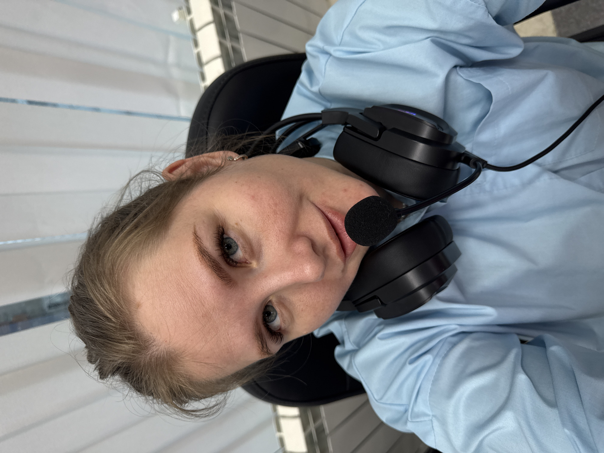 A young woman with light brown hair and blue eyes looks directly at the camera, showing a slight, friendly smile. She has fair skin, and her eyelashes appear prominent. She wears a light blue, collared shirt and has a black headset with a microphone resting around her neck. The microphone boom is positioned near her mouth. A small, silver hoop earring is visible on her left ear. She appears to be seated in a black chair, which is partially visible behind her shoulder. The background features horizontal elements, possibly blinds or a patterned wall, with light and darker stripes, suggesting an indoor setting like an office or home office. The overall impression is that she is pausing for a photo while potentially engaged in a work or communication activity. The image provides no specific time of day or weather cues. No visible text is present in the image. The location context provided is Perm, Russia.