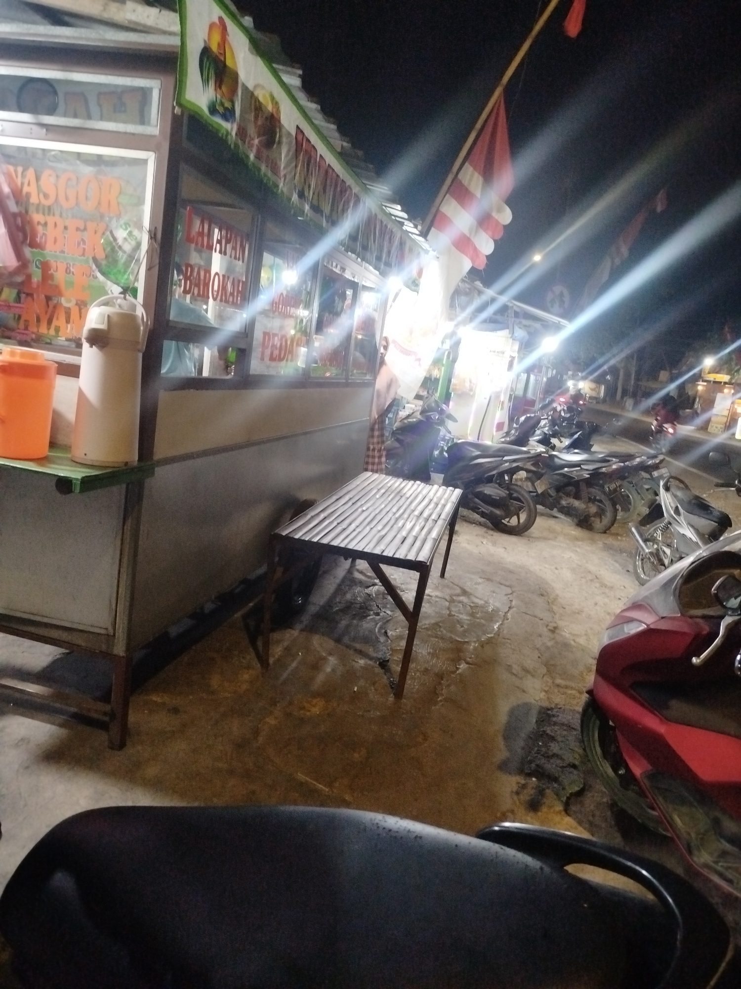 The image depicts a street scene at night, likely in Tonjung, Indonesia, with a food stall and parked motorcycles. The food stall, adorned with banners displaying text in Indonesian, appears to be selling "NASGOR SEBEK" and "LALAPAN BAROKAH." An orange container and a thermos are visible on a green-topped counter. A simple bamboo table and chairs are set up outside the stall. Several motorcycles are parked on the dirt and concrete ground in front of and beside the stall. A red motorcycle is prominently featured in the foreground. The atmosphere suggests a bustling local eatery.