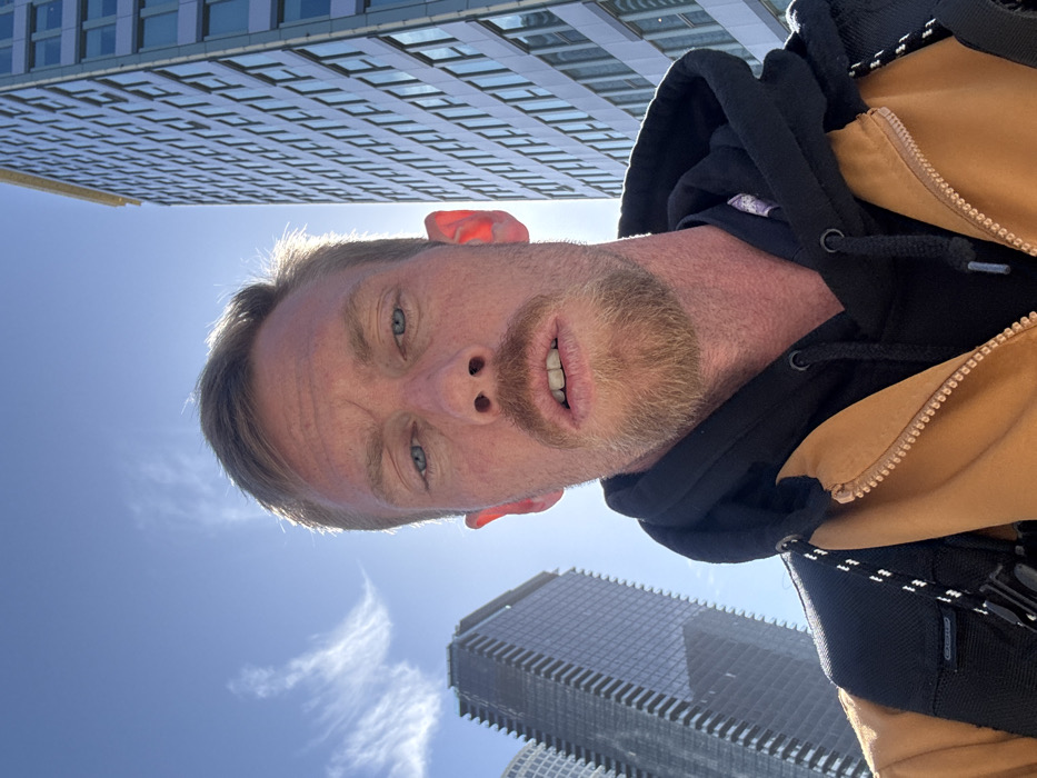 A selfie taken from a low angle features a light-skinned man with blond hair and beard stubble against a backdrop of skyscrapers and blue sky. He is wearing a black hoodie under a tan jacket. His eyes are blue. The sun is behind him, creating lens flare near his forehead and illuminating his ears. A large, modern building dominates the upper portion, while another smaller building is visible in the lower corner.