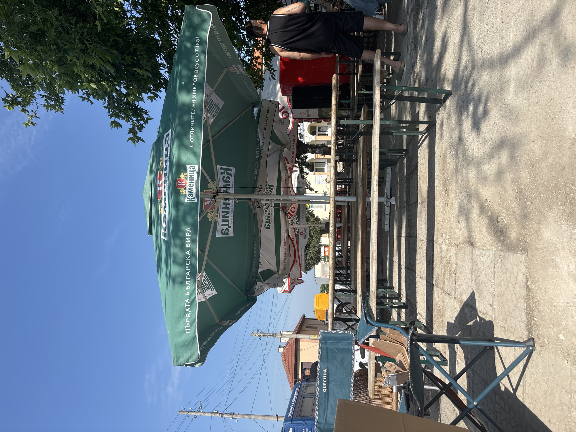 The image captures an outdoor seating area, likely a beer garden, in Kostievo, Bulgaria, on a clear, sunny day. The scene is dominated by multiple large green umbrellas, primarily one in the foreground, branded with "Каменица" (Kamenitza), a Bulgarian beer, featuring the slogans "ПЪРВАТА БЪЛГАРСКА БИРА" (The First Bulgarian Beer) and "С ОТЛИЧИТЕЛЕН ХМЕЛОВ ВКУС ОТ 1881" (With a distinctive hop taste from 1881).
Beneath the umbrellas, numerous wooden picnic tables with green metal frames are arranged in rows, mostly unoccupied. The ground consists of concrete paving slabs and dirt, with strong shadows cast by the umbrellas, tables, and nearby trees, indicating midday sun.
On the right side of the frame, a man stands with his back mostly to the viewer, wearing a black tank top and black shorts. Tattoos are visible on his left arm and leg, and he is wearing light-colored flip-flops. He appears to be observing the area to his left. Adjacent to him, a large red rectangular unit, possibly a cooler or vending machine, is present.
In the lower-left foreground, a blue folding camping chair, branded "QUECHUA," holds several paper bags. Next to it, part of a woven rattan chair is visible.
On the far left, a blue van with "Frühstück & Mittagstisch" partially visible on its side is parked. A man in a dark shirt is partially visible within the van's interior. Behind the blue van and several wooden utility poles with numerous overhead wires, a small building with a red tiled roof and light-colored walls is present. Further in the background, a yellow van is parked, and a larger building with a white facade and arched entrance is discernible. A large tree with green foliage is prominent on the right side, casting dappled shadows.