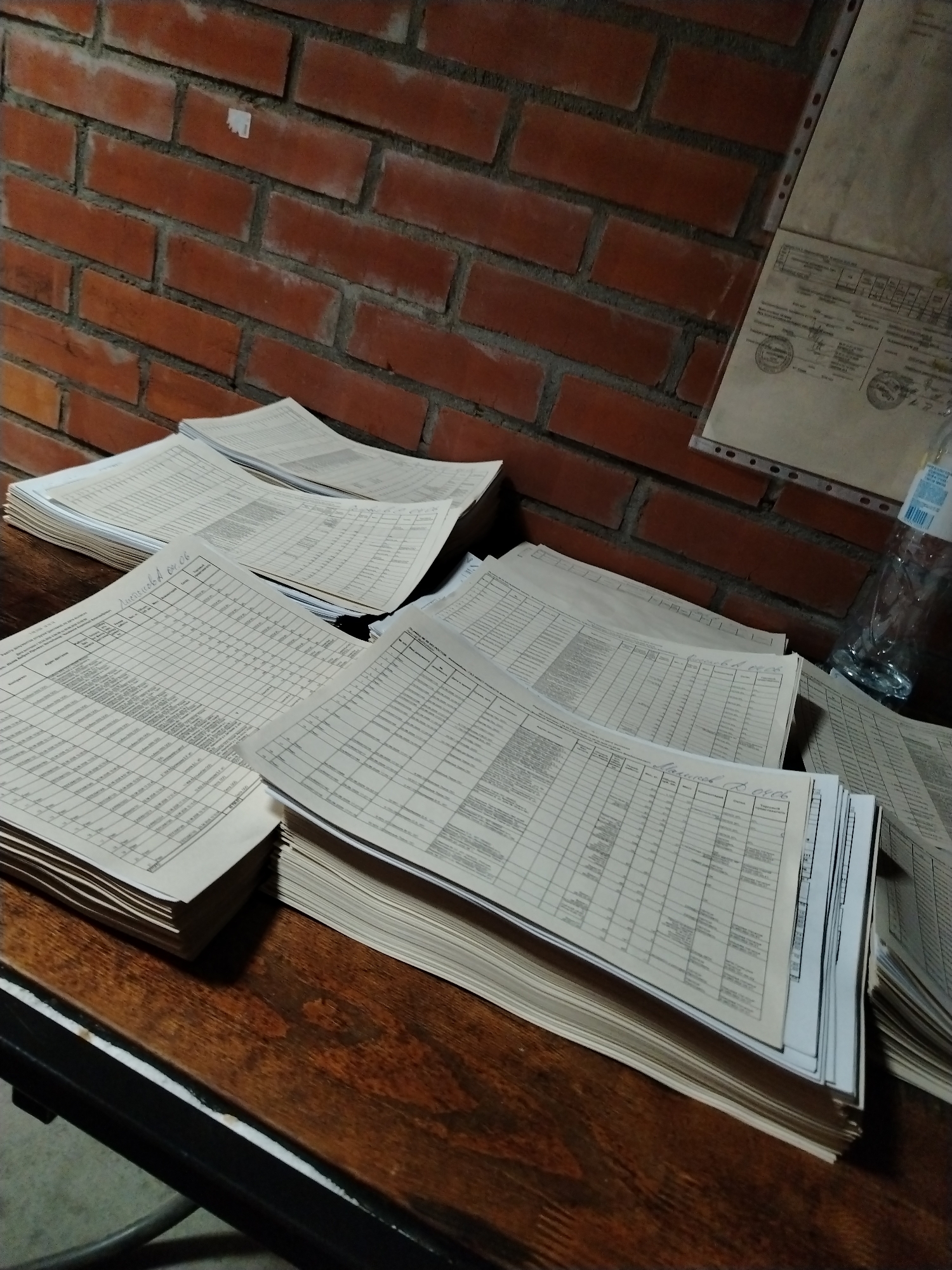 The image displays an indoor scene, likely a workspace or archive, dominated by numerous stacks of printed documents on a dark, richly-grained wooden table. A red brick wall serves as the background, suggesting an older building or an industrial-style interior.
The main subjects are the stacks of paper, which appear to be forms or spreadsheets filled with tabular data. Each sheet features a grid of columns and rows containing small, dense text, predominantly in Cyrillic script. Several stacks have handwritten annotations in blue ink on their top sheets. Visible handwritten text includes "ЛИТВИНОВА 08.06" (Litvinova 08.06) on one stack and "Матвеев В. 08.06" (Matveev V. 08.06) on another, possibly indicating names and dates. The large quantity of organized documents implies extensive record-keeping, administrative tasks, or data management.
On the brick wall in the upper right, a document in a clear plastic sleeve is visible, appearing to be an official form with Cyrillic text and what look like seals or stamps. A clear plastic water bottle is partially visible next to this hanging document.
The scene is illuminated by artificial lighting, which casts subtle shadows and gives the image a somewhat dim, focused ambiance. There are no people present, emphasizing the orderly arrangement of the paperwork. The setting is located in Шебунино, Russia.