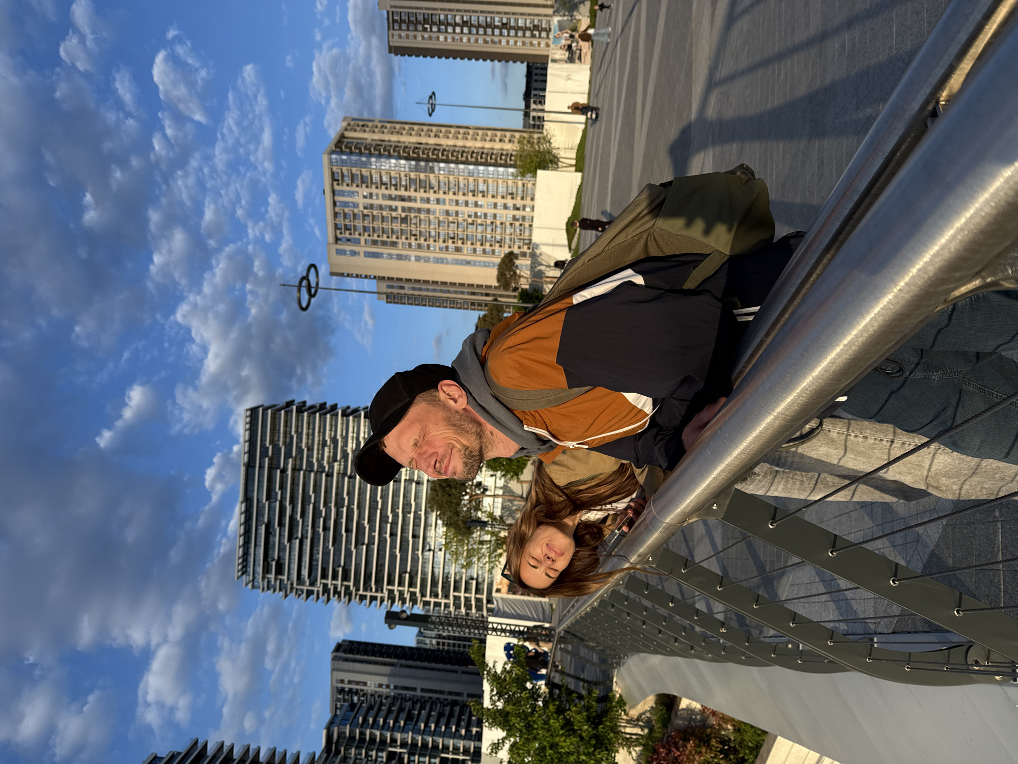 A rotated medium shot shows a man and a woman leaning over a metal railing, with high-rise buildings and a partly cloudy sky in the background. The man is wearing a dark baseball cap, a brown and black jacket, and has a beard. A green backpack rests on his back. The woman has long brown hair and is wearing a light-colored top. They appear to be looking down at something. The metal railing has horizontal cables running through it. The scene is located in Belgrade, Serbia, as evidenced by the architectural style of the buildings and the city's landmarks. The buildings are modern high-rises with geometric designs.