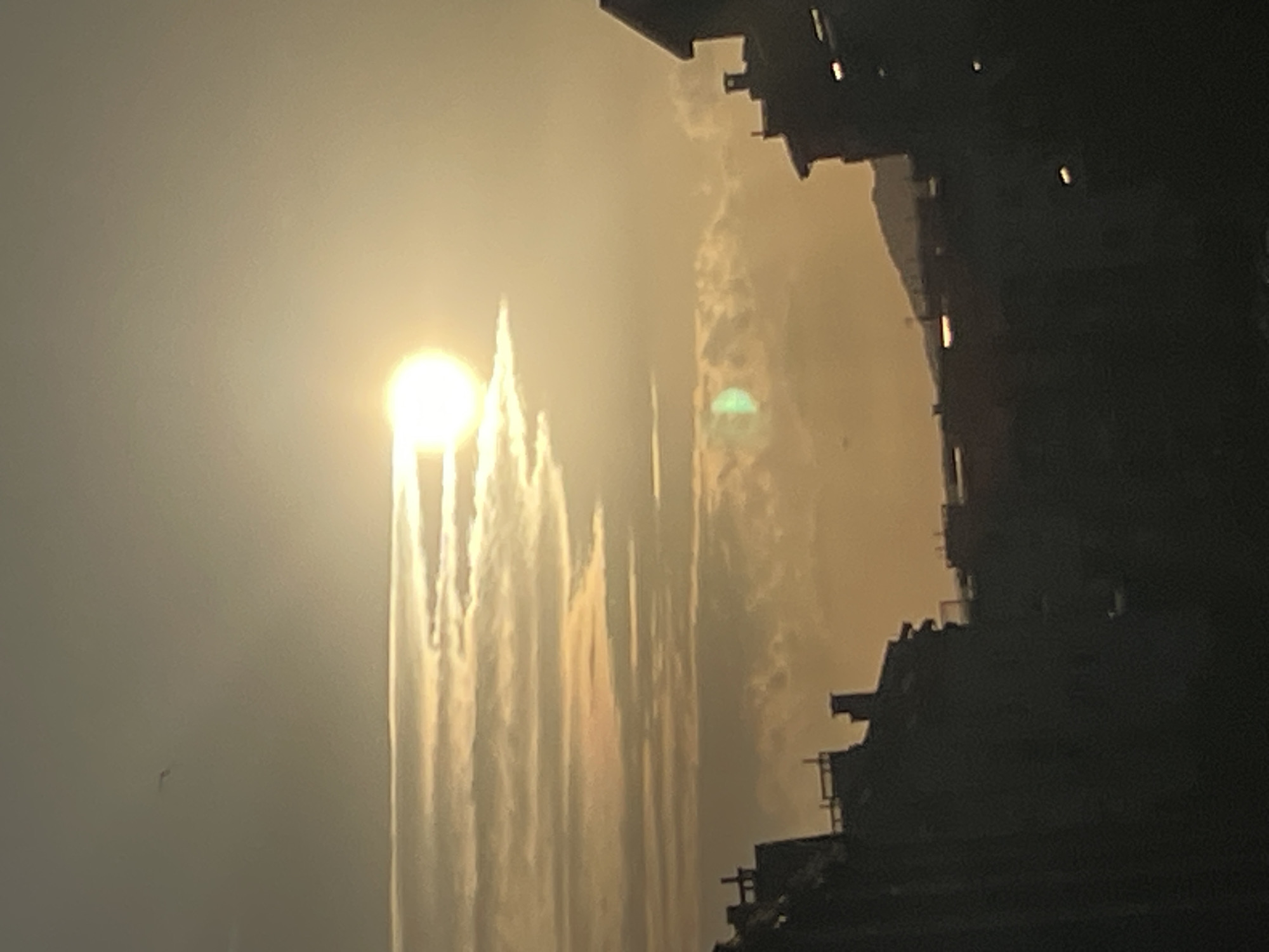 The image displays a striking sunset scene. The sun, a bright, luminous orb, is positioned in the upper left quadrant, casting a warm, golden glow across the sky. Tall, ethereal-looking structures, possibly a natural rock formation or man-made edifice, rise from the lower portion of the image, their forms softened by the atmospheric haze. The structures are silhouetted against the bright sky, with their edges appearing somewhat blurred.
To the right, a darker, more defined structure occupies the foreground, its silhouette creating a stark contrast with the luminous sky and the lighter formations. This structure appears to be man-made, possibly a building or a series of interconnected platforms, with visible levels and architectural details, though they are rendered in shadow.
The sky is filled with soft, diffused light, suggesting either mist, fog, or heavy clouds that are illuminated by the setting sun. Wispy streaks of light emanate from behind the lighter, central structures, creating a dramatic effect. There is a hint of green light, possibly a lens flare, within the illuminated mist to the right of the central structures. The overall impression is one of a dramatic and somewhat mysterious natural or urban landscape at dusk. No text is visible in the image.