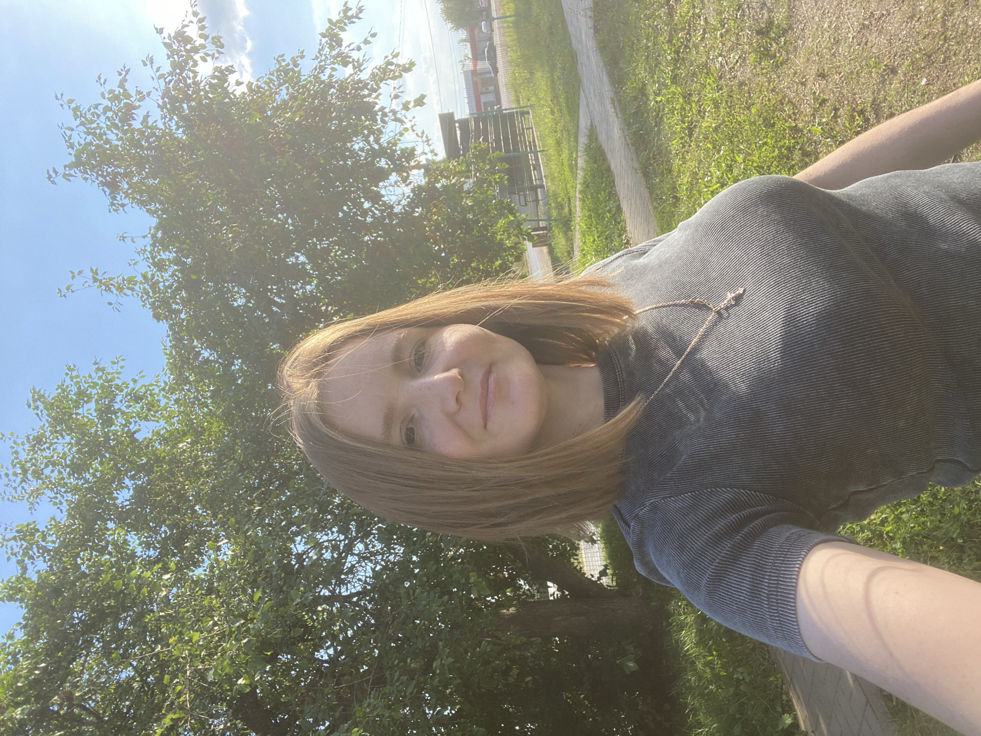 A young woman with shoulder-length reddish-brown hair and fair skin, wearing a dark gray ribbed t-shirt and a delicate gold chain with a cross pendant, is taking a selfie outdoors in Chaikovsky, Russia. The sun is shining brightly, casting a warm glow on the scene. Behind her, a large, leafy tree with a thick trunk stands to the left. To the right, a paved walkway runs parallel to a grassy area. In the background, there is a building structure with fencing and a road with passing cars. The sky is blue with some clouds. The woman is smiling slightly at the camera.
