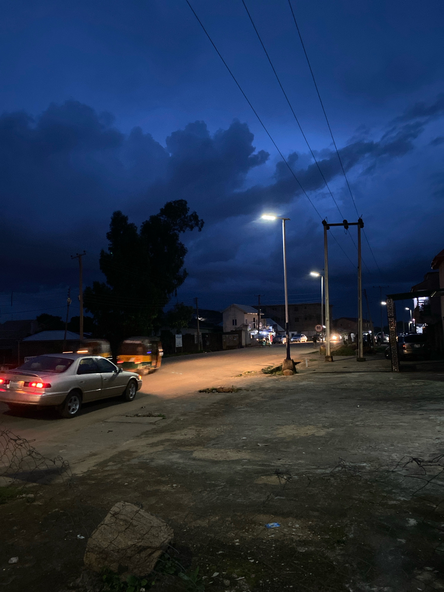 The image captures a street scene in Jos, Nigeria, during twilight or early evening. The sky is a deep, stormy blue, filled with dark, textured clouds. Several utility poles with power lines stretch across the upper portion of the frame. A prominent streetlamp, illuminated with a bright white light, stands on the left side of the road, casting a warm glow on the pavement.
In the foreground, a light-colored sedan is parked or slowly moving, its taillights illuminated. Behind it, a yellow and green motorized rickshaw, often seen in Nigerian cities, is visible, its headlights also on. The road surface appears to be a mixture of asphalt and dirt, with some debris and scattered rocks visible. On the right side of the road, there are buildings with windows and some architectural details, including what looks like an arched entrance. Another streetlamp is visible further down the road, and people can be seen in the distance, some gathered near buildings or walking. A speed limit sign indicating "50" is mounted on a pole along the street. The overall atmosphere is dusky and somewhat moody, typical of the transition from day to night, possibly with impending rain due to the dark clouds.