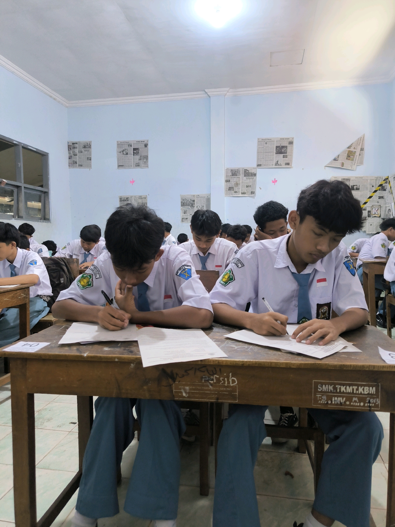 The image depicts a classroom scene at SMK.TKMT.KBM, identified by writing on the wooden desk, possibly located in Kebumen, Indonesia. Several male students, dressed in white shirts with blue ties and blue pants, are seated at wooden desks, actively engaged in taking a test or exam. Each student has papers and a pen in front of them. The classroom walls are painted a light blue and are adorned with various newspaper clippings. The lighting appears to be artificial, emanating from the ceiling. The overall atmosphere suggests a formal academic setting.