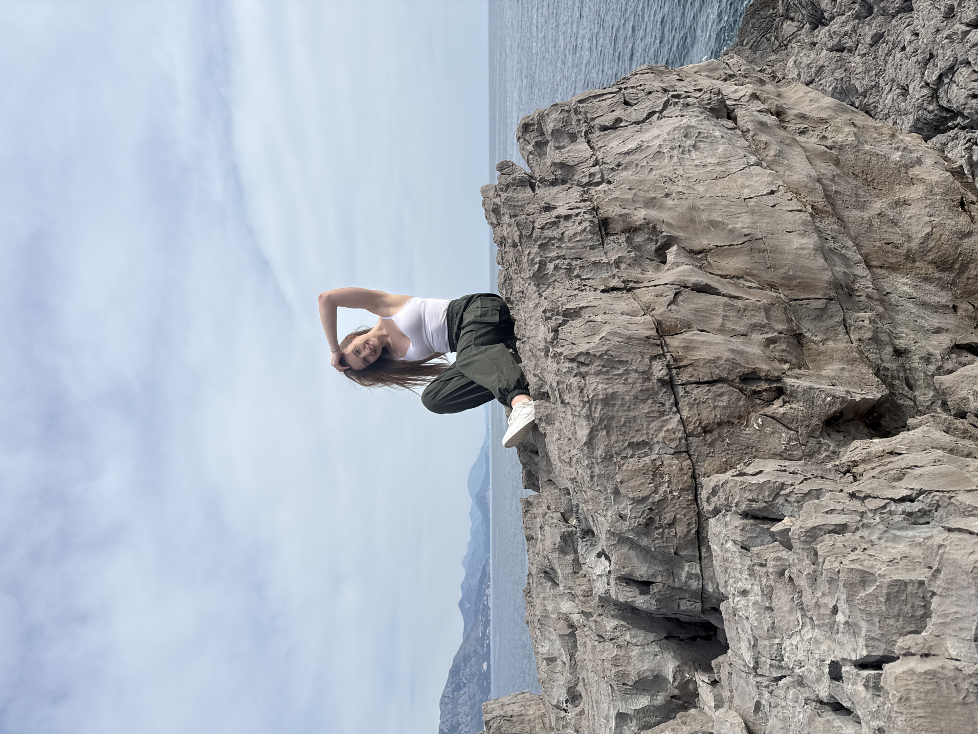A woman is perched on a rocky cliff overlooking the Adriatic Sea near Boreti, Montenegro. She wears a white tank top and dark green cargo pants with white sneakers. Her long, brown hair falls down her back as she poses with one arm raised above her head. The cliff face is composed of gray rock formations with the sea visible in the background. The sky is overcast.