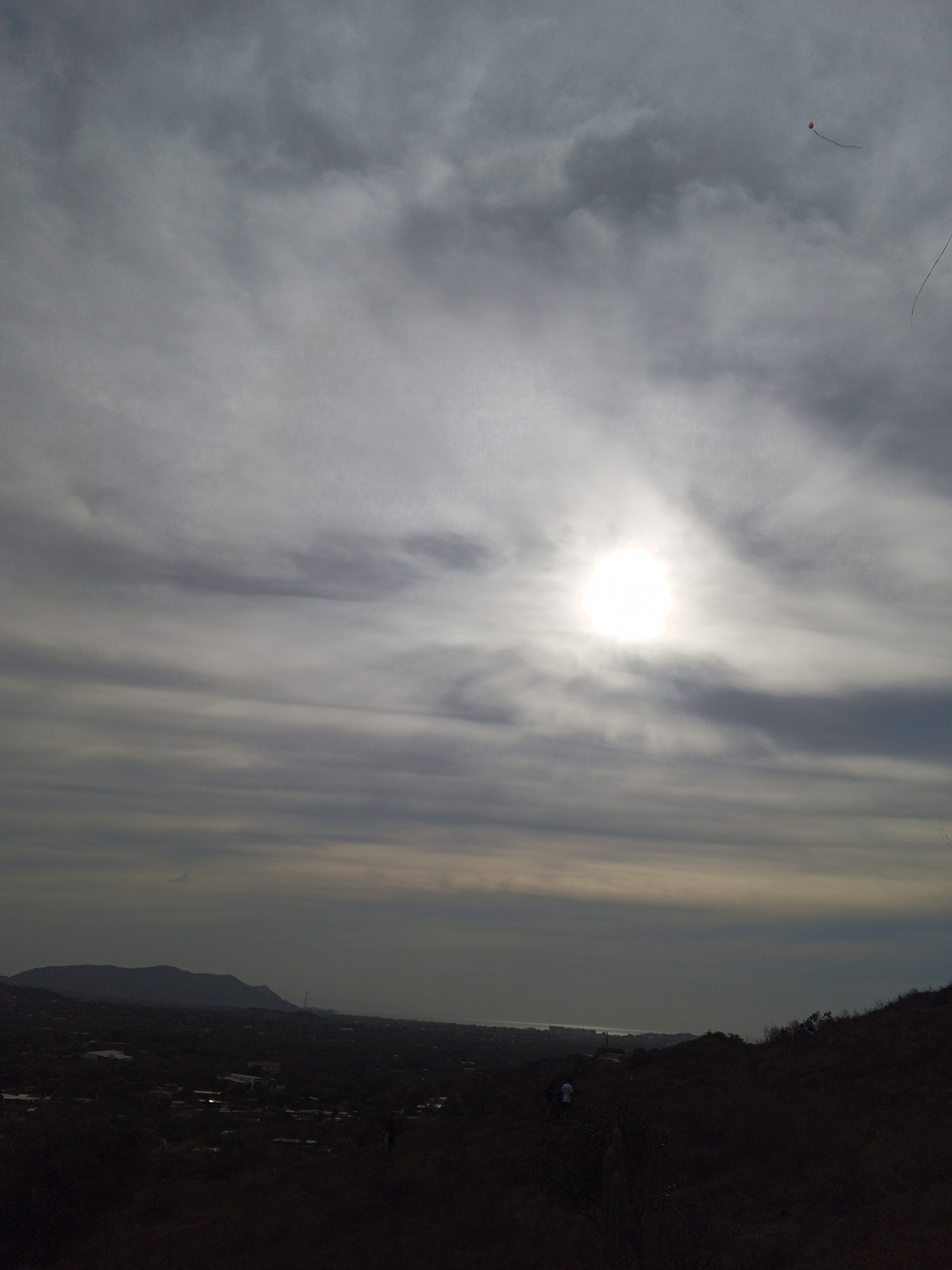 The image shows a landscape with a cloudy sky and the sun obscured by the clouds. In the upper right, a small kite is visible. Below the horizon, the landscape slopes down to a distant urban area, with hills visible in the far background. The foreground is dark, likely a hillside or ridge, with vegetation and possibly some people walking on it.