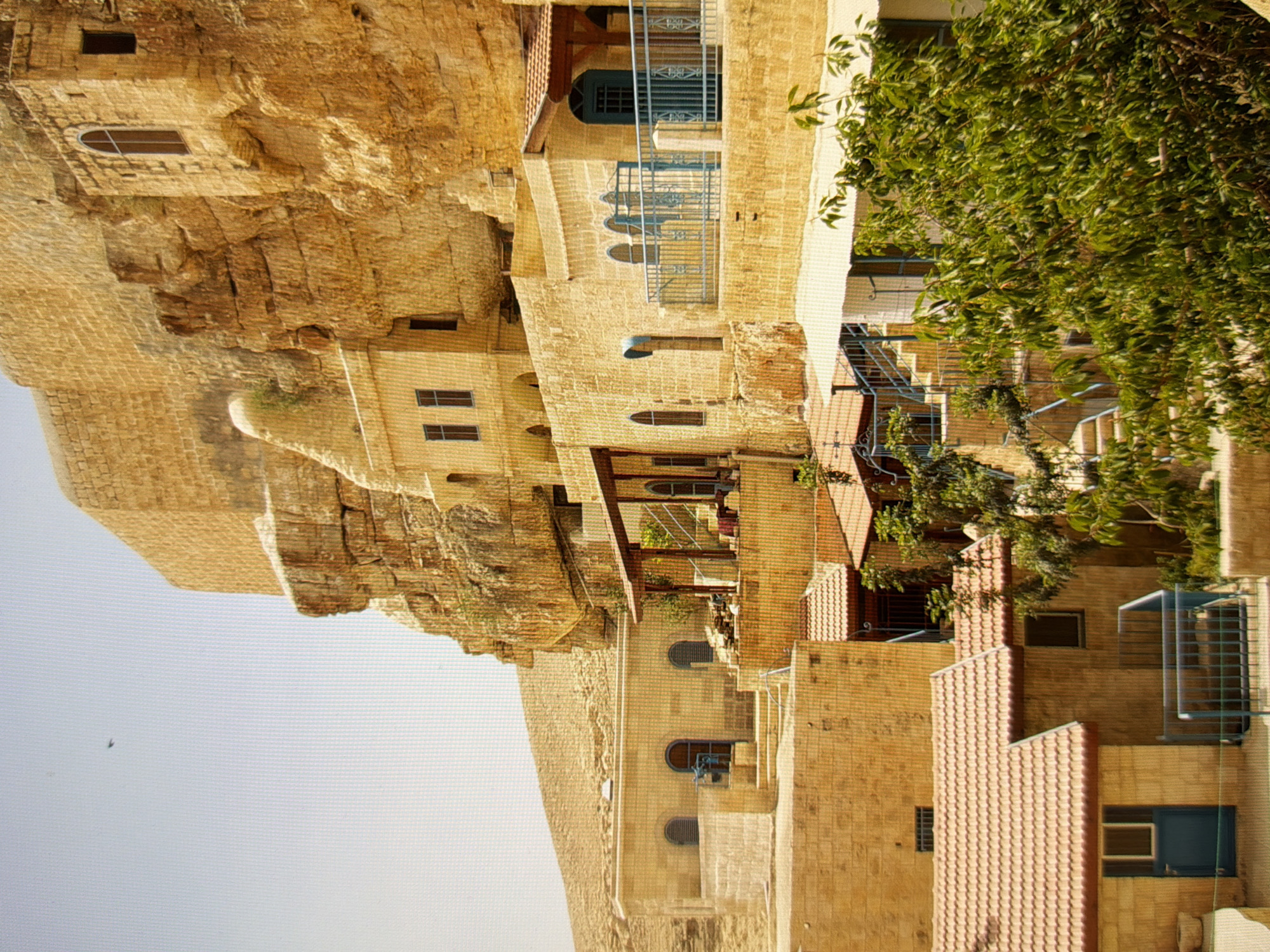 The image depicts a complex of stone buildings constructed into a rocky hillside. The architecture is characterized by the use of light-colored stone, arched doorways and windows, and red-tiled roofs. Several levels of structures are visible, some built directly into the rock face, while others are freestanding but integrated with the natural terrain. Blue railings and some blue-painted doors and windows add accents to the predominantly earthy tones. Green foliage from trees and bushes is present in the foreground and interspersed with the buildings. The sky is a pale blue. The scene is one of an ancient settlement or monastery, likely in a dry, mountainous region. This image is not located in Kyiv, Ukraine.