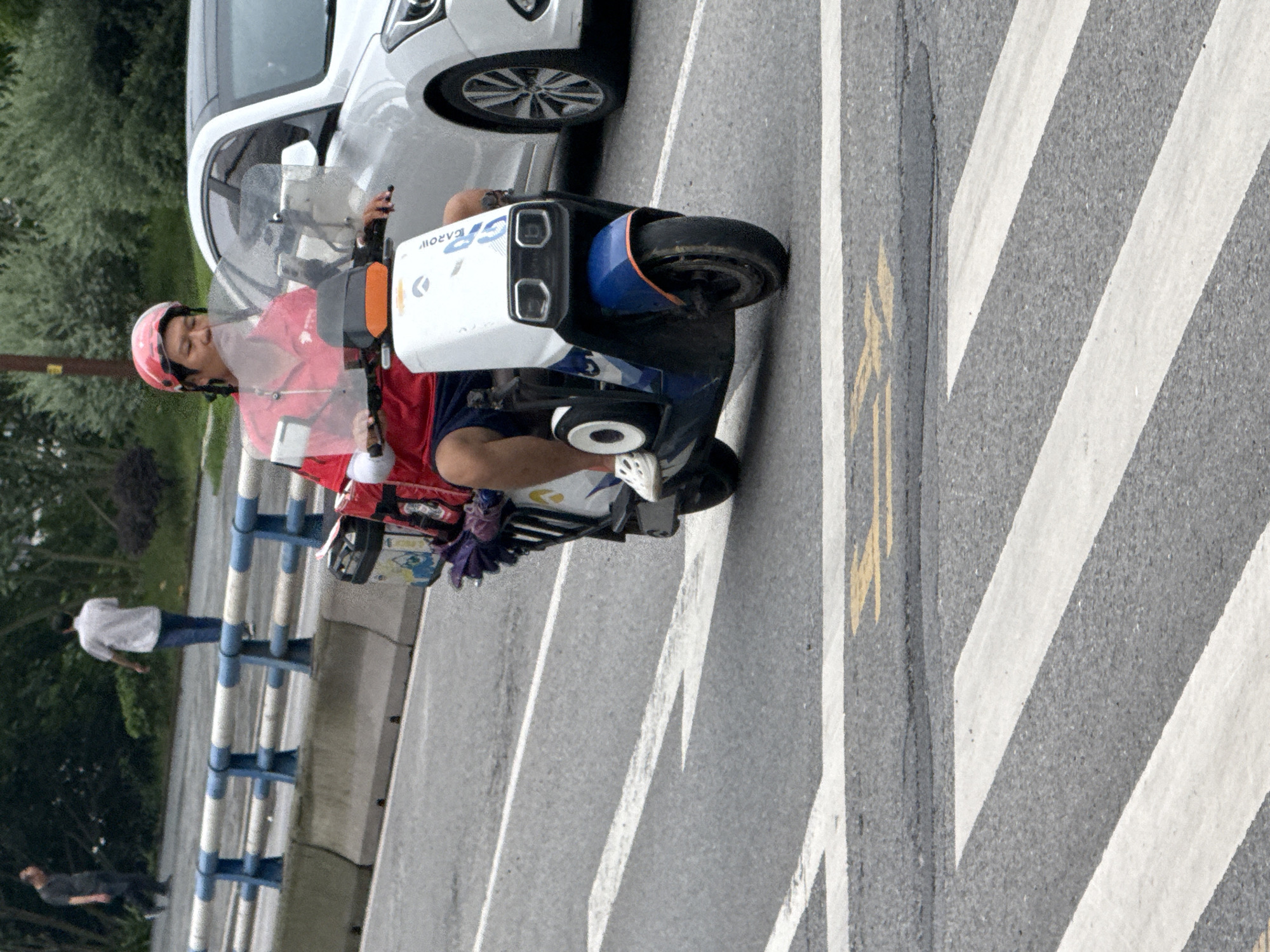 The image captures a street scene in Shapingba District, China, focusing on a person riding a white and blue electric scooter. The rider, wearing a red helmet and a red shirt with white arm coverings, is positioned in the middle of a road with visible white lane markings. A white car is visible to the right of the scooter. In the background, two pedestrians are walking on a sidewalk separated by a concrete barrier and a blue and white railing. The environment appears to be urban, with some greenery visible behind the railing. The scooter has a storage compartment at the back.