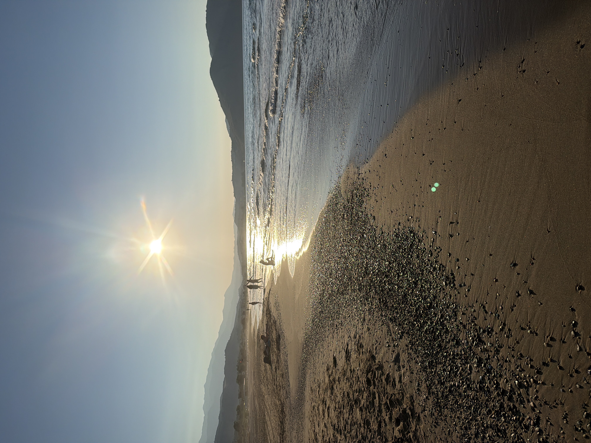 This image depicts a sunset scene on a beach in Georgioupoli, Greece. The sun is low in the sky, casting a bright glare and long shadows across the sand and water. Gentle waves are lapping at the shore, creating a shimmering reflection of the sunlight. Several people are visible on the beach, some walking and others sitting by the water's edge. In the background, mountains form a silhouette against the horizon. The beach itself appears to be a mix of sand and small pebbles.