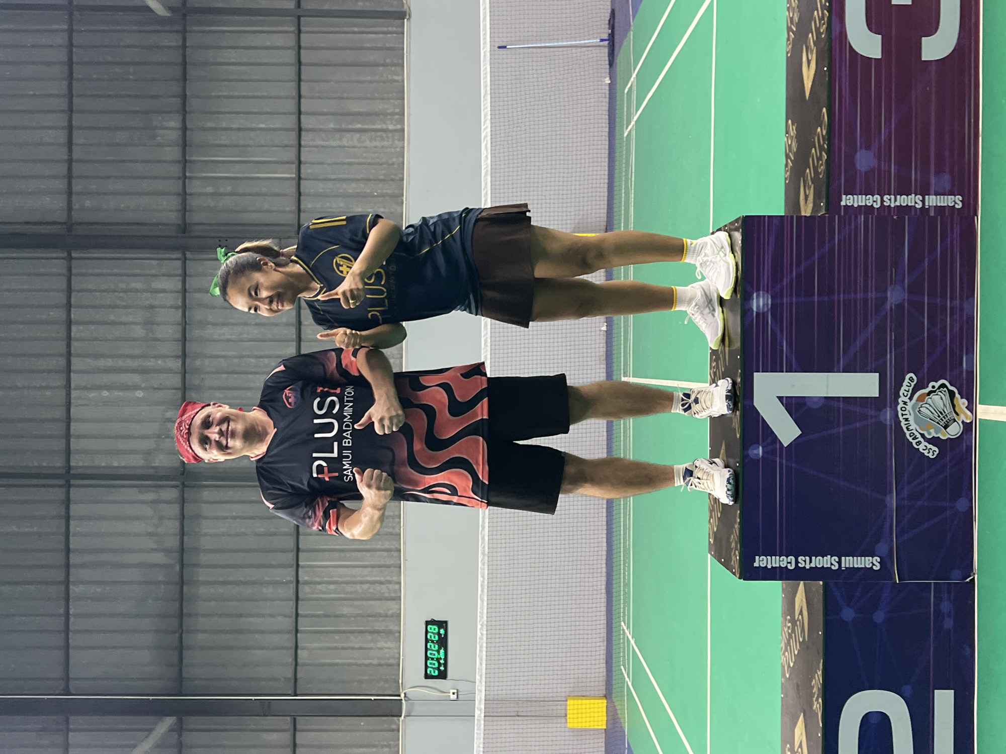 Two individuals, a man and a woman, are standing on a winner's podium in what appears to be an indoor badminton court. The man, on the left, is wearing a black and red patterned t-shirt with the text "PLUS1 SAMUI BADMINTON" and a red bandana. He is giving a thumbs-up with both hands. The woman, on the right, is wearing a dark blue t-shirt with yellow stripes and the "PLUS1" logo, and a brown skirt. She is also giving a thumbs-up with both hands and has a green hair tie. They are both wearing athletic shoes. The podium is decorated with graphics including the number "1" prominently displayed on the left side. The background shows a badminton net, court markings, a digital clock displaying "20:08:28", and a corrugated metal wall. The overall setting is consistent with a sports facility in Ko Samui District, Thailand, likely the Samui Sports Center.