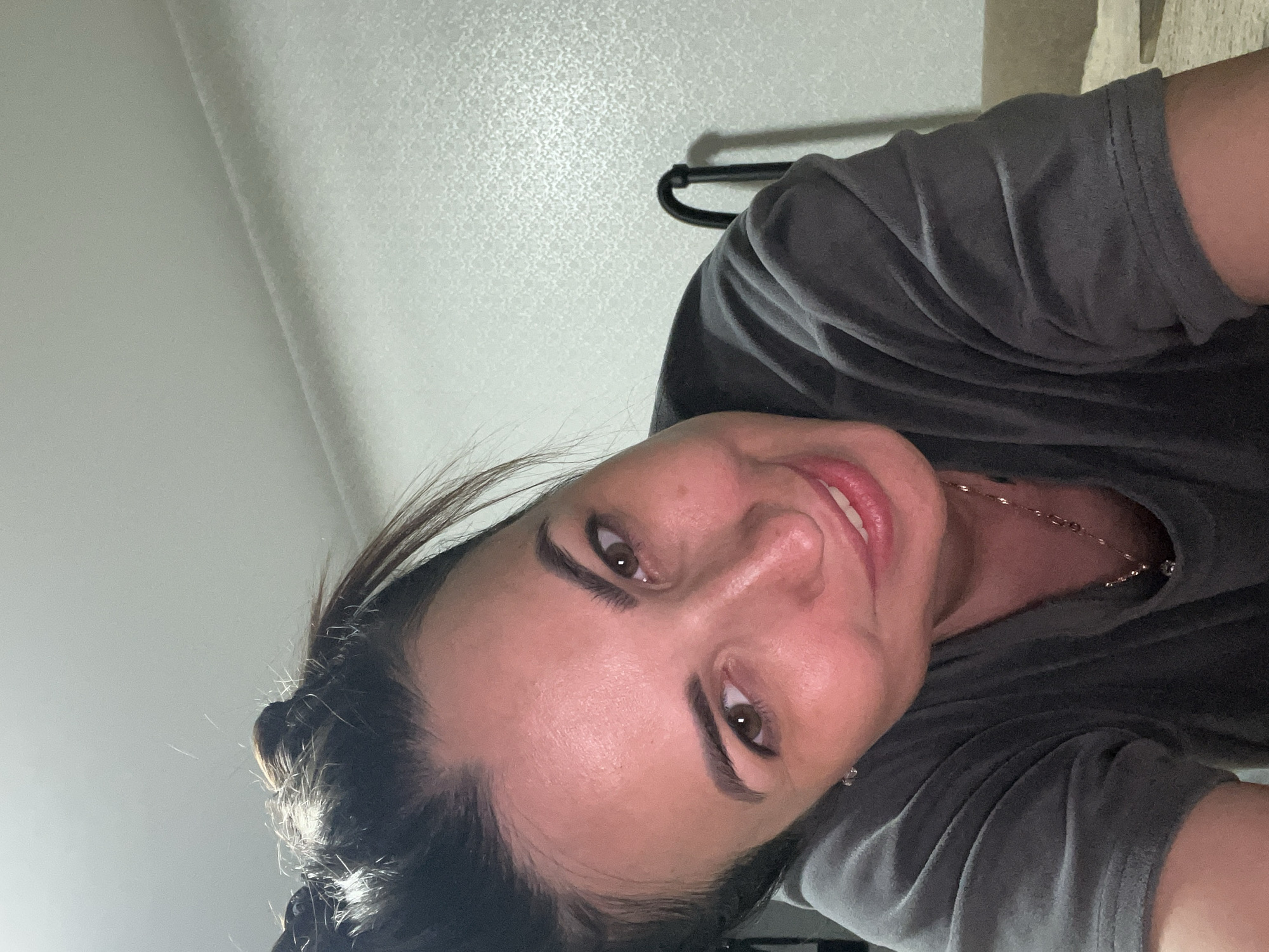 This is a close-up, self-portrait of a woman, taken from a low angle, with the image rotated 90 degrees counterclockwise. The woman has dark hair pulled back, with strands framing her face. She is wearing a dark gray t-shirt and a delicate necklace. Her eyes are brown and she is looking directly at the camera with a slight smile. Her makeup appears to include eyeliner and mascara. The background is a textured, light-colored wall, with a hint of a coat hanger or similar object visible in the upper right portion of the frame. The lighting suggests an indoor setting, possibly at night or in a room with artificial light. The overall mood conveyed is casual and friendly. There is no visible text in the image.