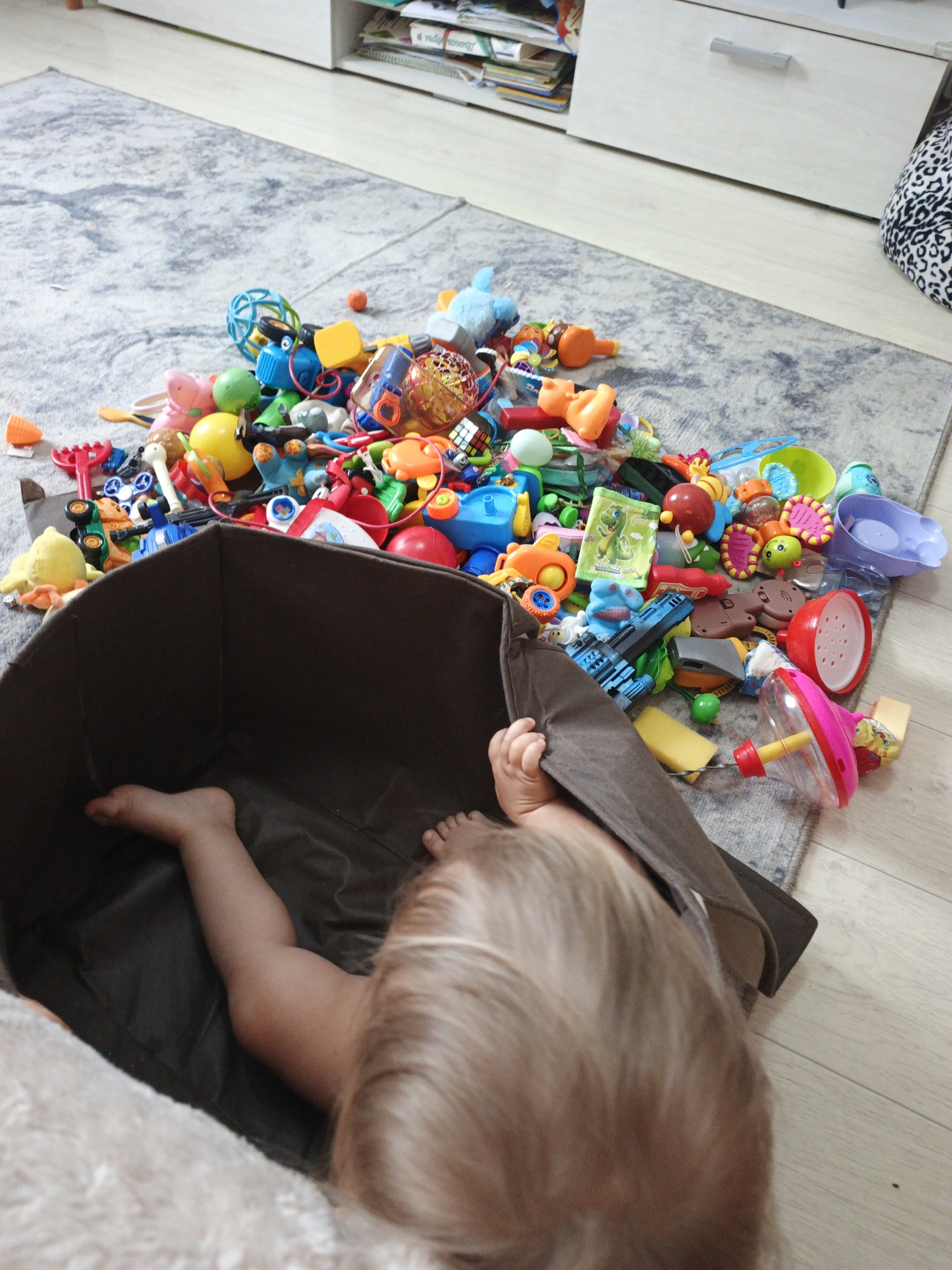 An infant is partially visible, lying on its side within a dark brown fabric storage bin. The infant's legs are extended and visible within the bin, and their back and head are facing away from the camera. The bin is overflowing with a large assortment of colorful children's toys, which are scattered across a gray and white patterned rug. The toys include building blocks, plastic animals, toy vehicles, spinning tops, and other miscellaneous play items. The rug is placed on a light-colored wooden floor. In the background, a white cabinet with shelves filled with books and other items is visible. The scene appears to be an indoor play area or living room. The image was taken in Zlatoust, Russia.