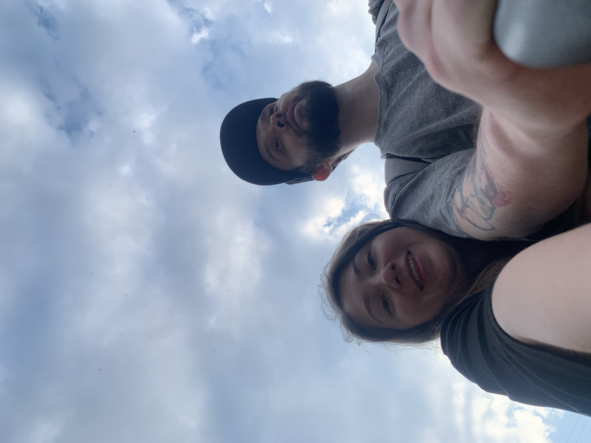 A low-angle selfie is taken by two individuals against a cloudy sky, with the camera positioned below eye level. The person in the foreground on the left is a woman with dark hair and a smiling expression, wearing a black t-shirt. To her right, a man with a beard and a black baseball cap is looking upwards. He is wearing a grey t-shirt. His right arm is visible in the foreground, resting on a grey object. Tattoos are visible on his arm. The background consists of a cloudy sky with some small, dark specks that could be insects or birds. There is no indication of the city of Chaikovsky, Russia, or any other specific location.