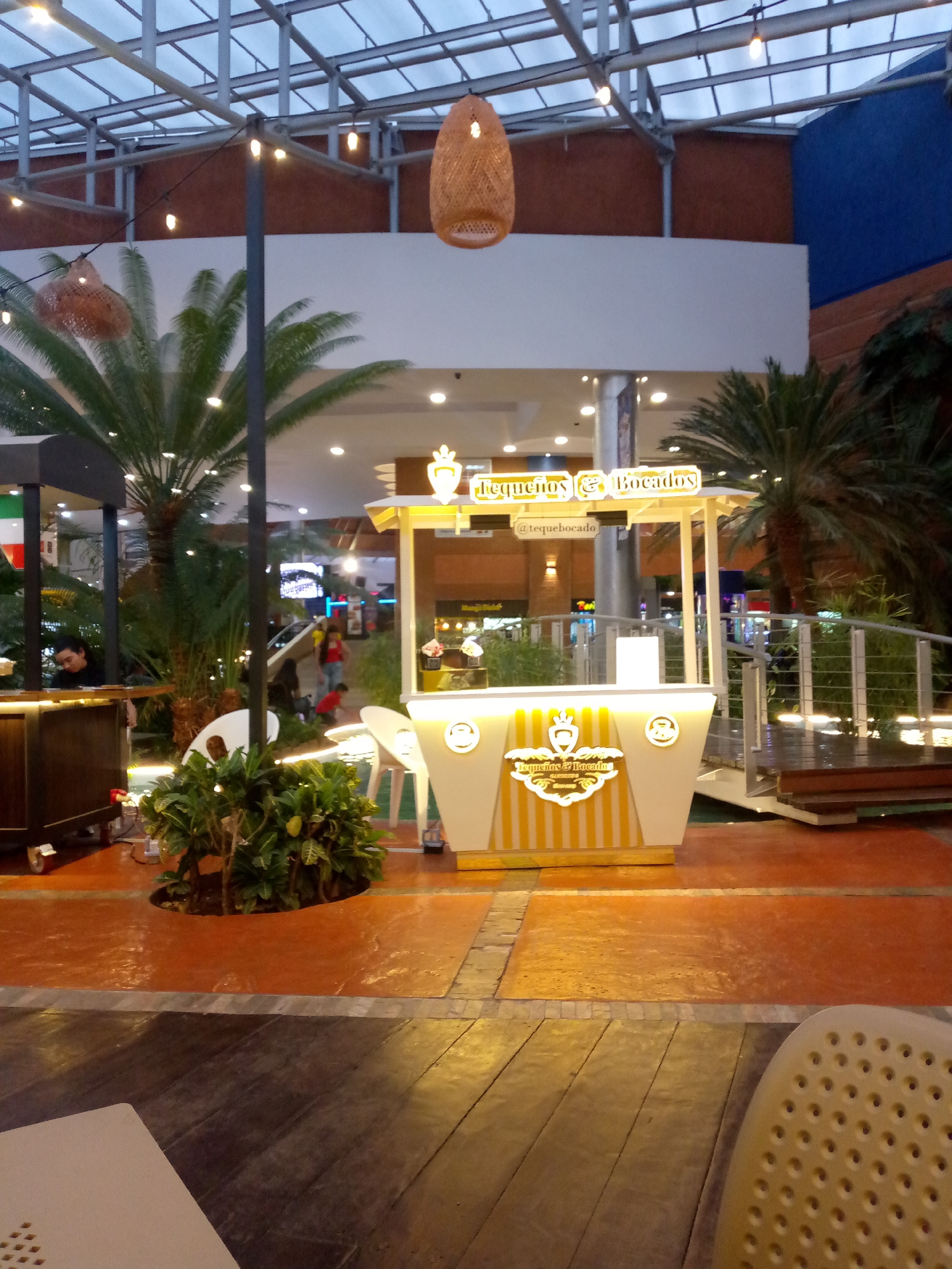 The image shows an indoor scene in what appears to be a shopping mall or large public space. The flooring is a mix of orange-toned tiles and wooden planks, creating a textured surface. The scene is dominated by a food kiosk identified by the sign "Tequeños & Bocados." The kiosk is decorated in yellow and white stripes and is well-lit. Lush greenery, including palm trees and various plants, is scattered throughout the space, contributing to an open and airy feel. Additional kiosks are visible in the background, with one partially obscured on the left. Overhead, the lighting fixtures and the mall's architectural details are noticeable, including a woven lampshade and a structured ceiling. A metal and wooden bridge is visible near the main kiosk. People can be seen walking and interacting in the background.
