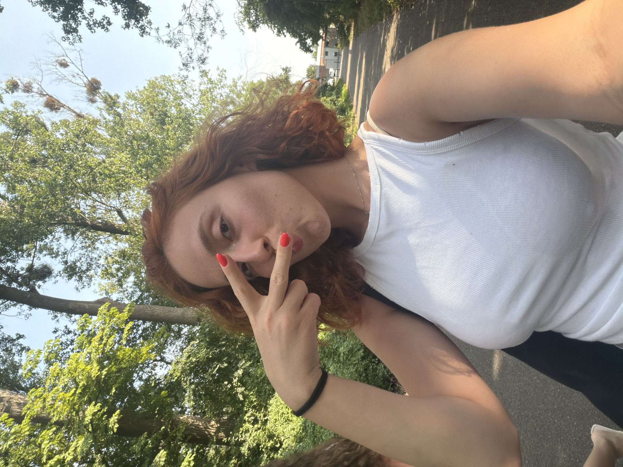 In this selfie taken outdoors, a young woman with wavy red hair and bright red nail polish is the central subject. She is wearing a white ribbed tank top and a silver necklace. Her right hand is held up to her face, making a peace sign with her index and middle fingers, while her lips are puckered. She is facing the camera directly, with the background featuring a tree-lined pathway. The path appears to be a road or sidewalk, with some buildings visible in the distance and cars parked along its side. The trees are lush and green, indicating it's likely daytime with sunlight filtering through the leaves. This scene is set in Illkirch-Graffenstaden, France.