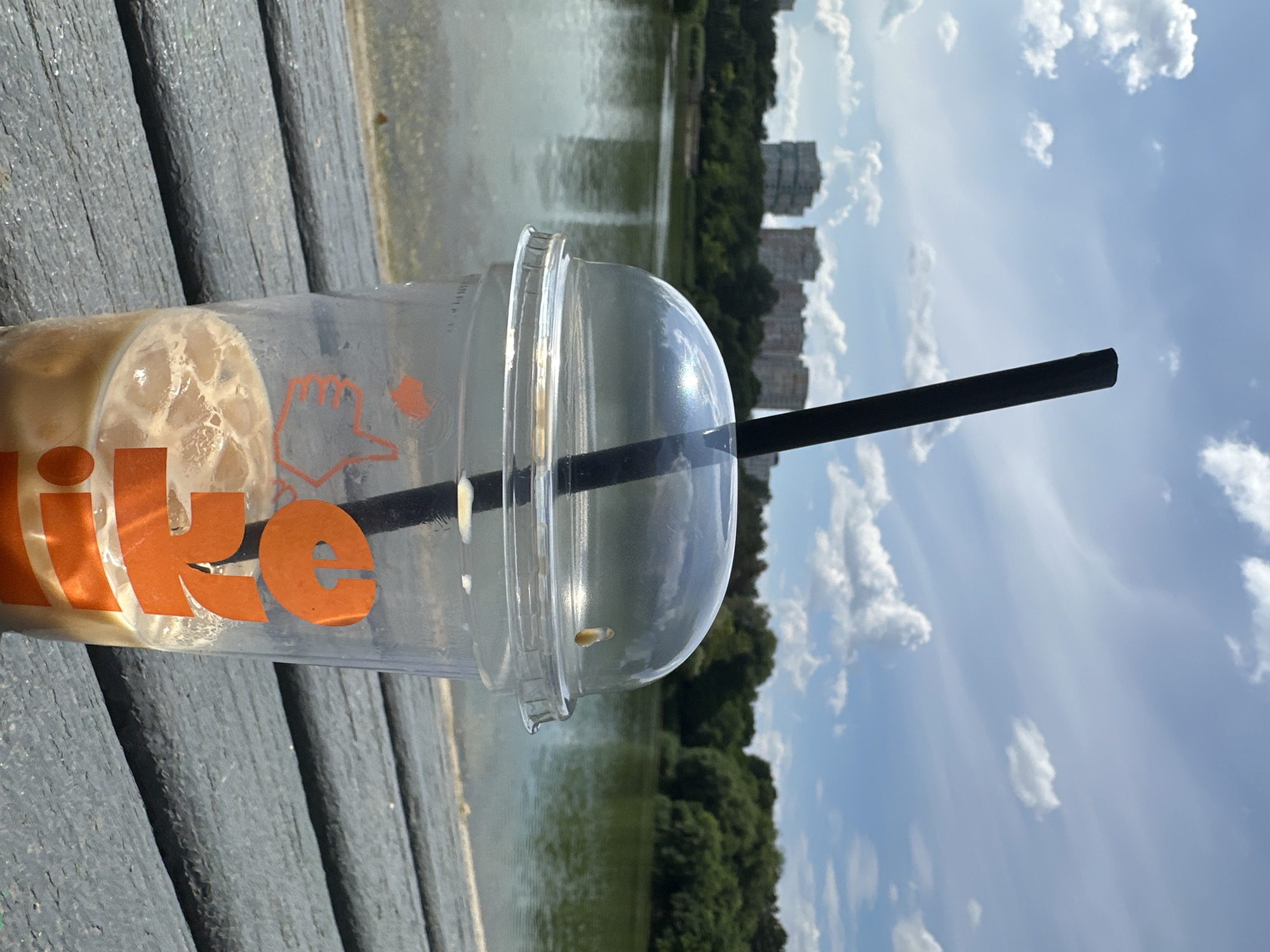 A close-up shot focuses on a plastic cup with a clear dome lid and a black straw, containing an iced coffee drink with ice cubes and a creamy beverage. The cup features orange lettering that appears to spell out "JOY" and a thumb-up graphic. It rests on a weathered wooden surface, possibly a table or deck. In the background, a body of water, likely a lake or river, stretches out, reflecting the sky. Across the water, a line of lush green trees forms a natural border, behind which are several multi-story buildings. The sky above is a clear blue with scattered white cumulus clouds. The scene suggests an outdoor recreational area within Chișinău, Moldova, possibly a park or waterfront, on a bright, sunny day.