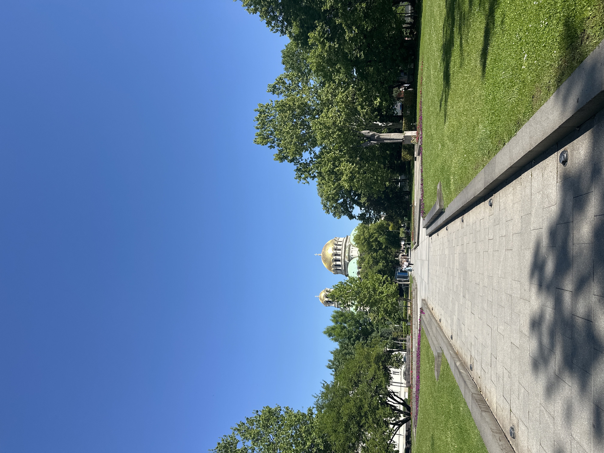 The image shows a view towards Alexander Nevsky Cathedral in Sofia, Bulgaria. The cathedral's golden domes are visible through the trees lining a paved walkway. The sky is clear and blue. On the right side of the walkway is a strip of green grass. There are people in the distance.