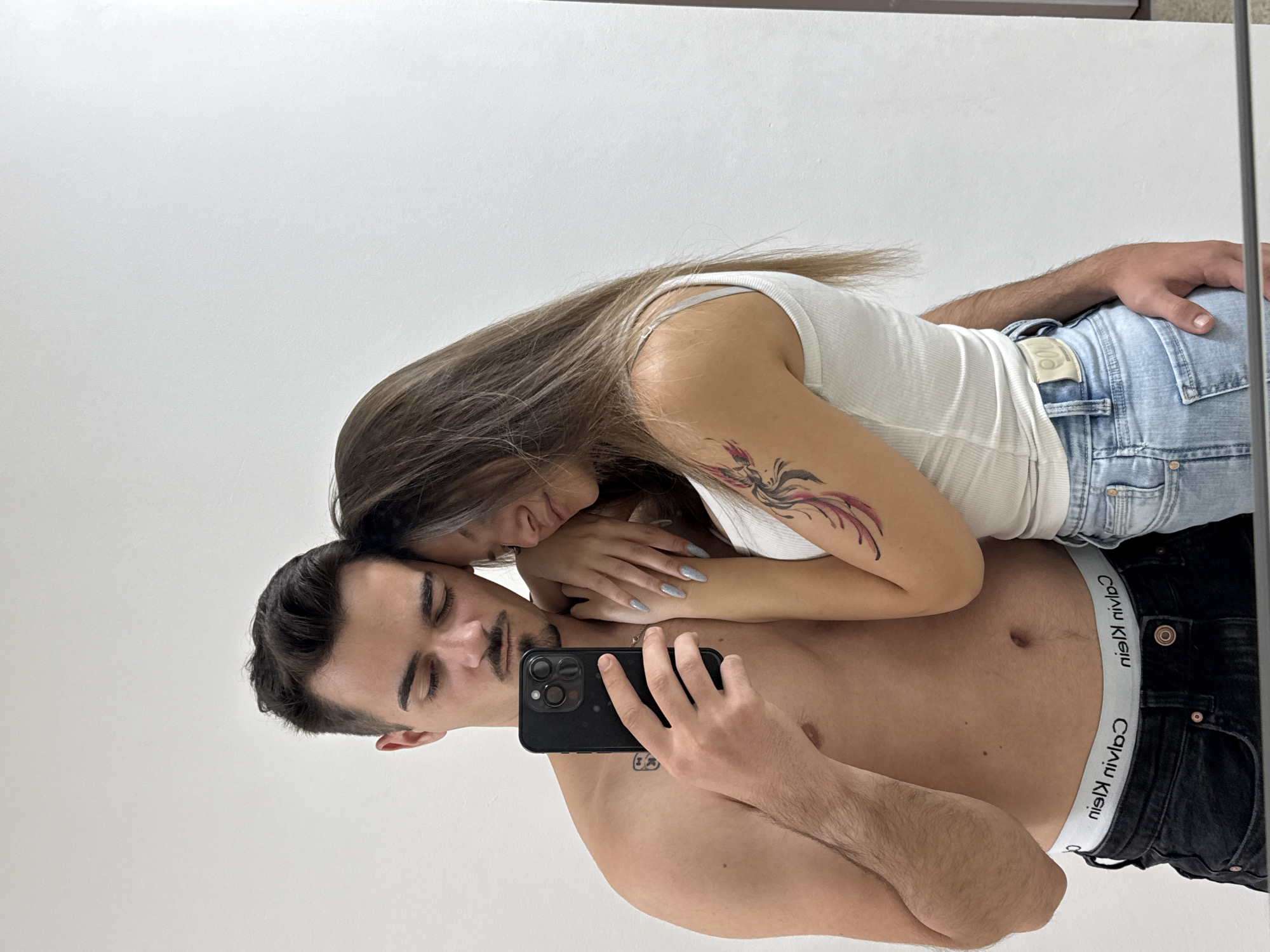 The image is a mirror selfie depicting two individuals, a male and a female, embraced in an indoor setting against a plain white wall.
The male, appearing to be in his early to mid-20s, has dark brown hair, a mustache, and a goatee. He is bare-chested, wearing black denim jeans and white Calvin Klein boxer briefs, with the reversed "Calvin Klein" branding visible on the waistband. A small, dark tattoo is visible on his left chest/shoulder area. He holds a black smartphone, featuring three prominent camera lenses and a flash, in his right hand, angled to capture the reflection. His left arm is positioned around the female.
The female, also appearing to be in her early to mid-20s, has long, light brown hair. Her head is leaning against the male's right shoulder, with her eyes closed or directed downwards. She wears a white ribbed tank top and light blue denim jeans. A multi-colored tattoo, depicting a feathered design in red, black, purple, yellow, and white, is visible on her upper left arm. Her right arm is around the male's waist/hip, and her left hand rests on his shoulder. Her fingernails are long and coated in a light grey/silver glitter polish.
Both individuals have their eyes closed or are looking downwards. The background is a stark white wall. The bottom edge of the mirror is visible, showing a slight reflection of the floor. There are no discernible visual cues within the image to specify the location as Sofia, Bulgaria, or any other particular city or country.