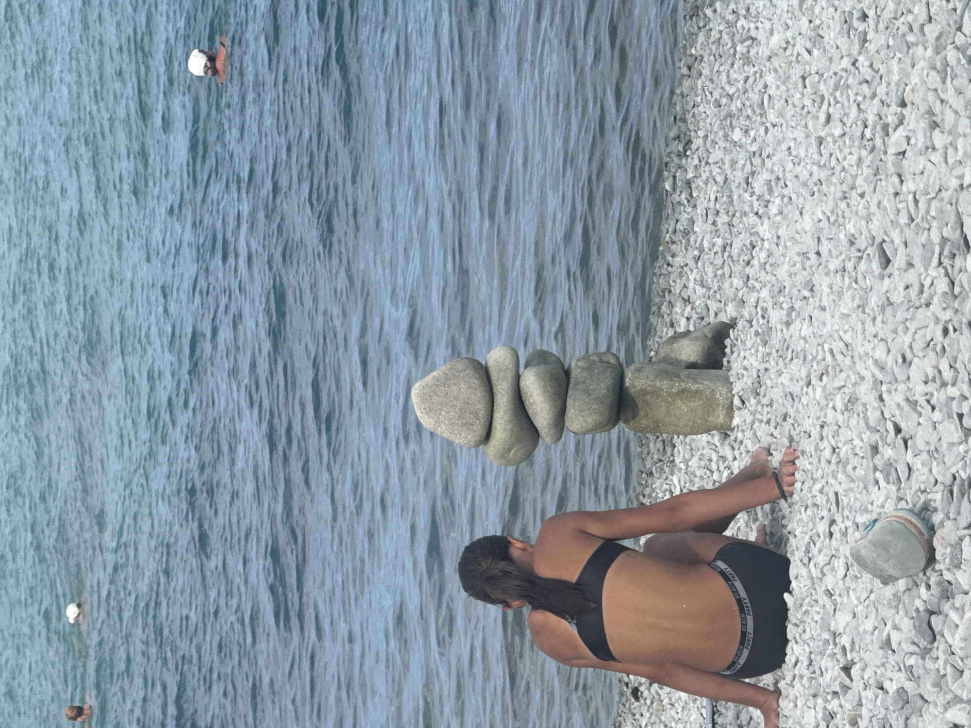 A vertical, slightly low-angle shot captures a serene beach scene with a woman in the foreground and a precarious stone sculpture in the middle ground. The woman, facing away from the camera, is seated on a pebble-strewn shore, her back to the viewer. She wears a black bikini with a band that reads "BEACH PARTY" in white text. Her dark hair is pulled back, and her arms are resting on her knees. To her left, a stacked arrangement of smooth, grey stones rises from the water. The largest stone at the top is a rough, rectangular piece. The water is a calm, blue expanse with gentle ripples. In the distance, a few people are visible in the water, one with a white hat. The sun appears to be high in the sky, casting soft light. The location is Partenit, Ukraine.
