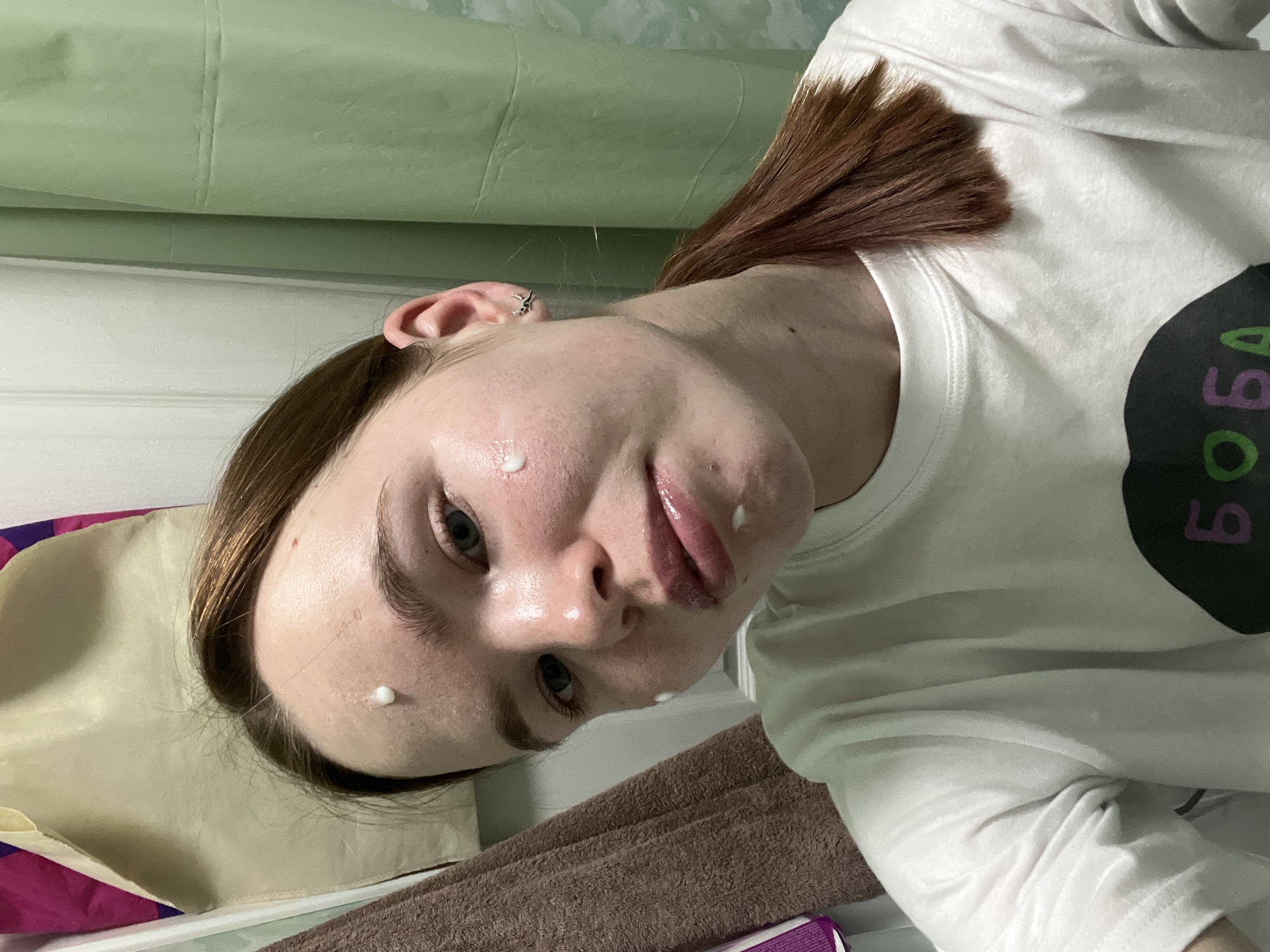 A close-up shot depicts a young woman with pale skin, a few freckles, and light blue eyes. There are small dabs of white cream visible on her forehead, cheek, and chin. Her reddish-brown hair is pulled back, and she is wearing a white t-shirt with a design on it. The background is indistinct, but suggests a domestic interior. Based on the T-shirt logo, it can be assumed that the photograph was taken in Chaikovsky, Russia.