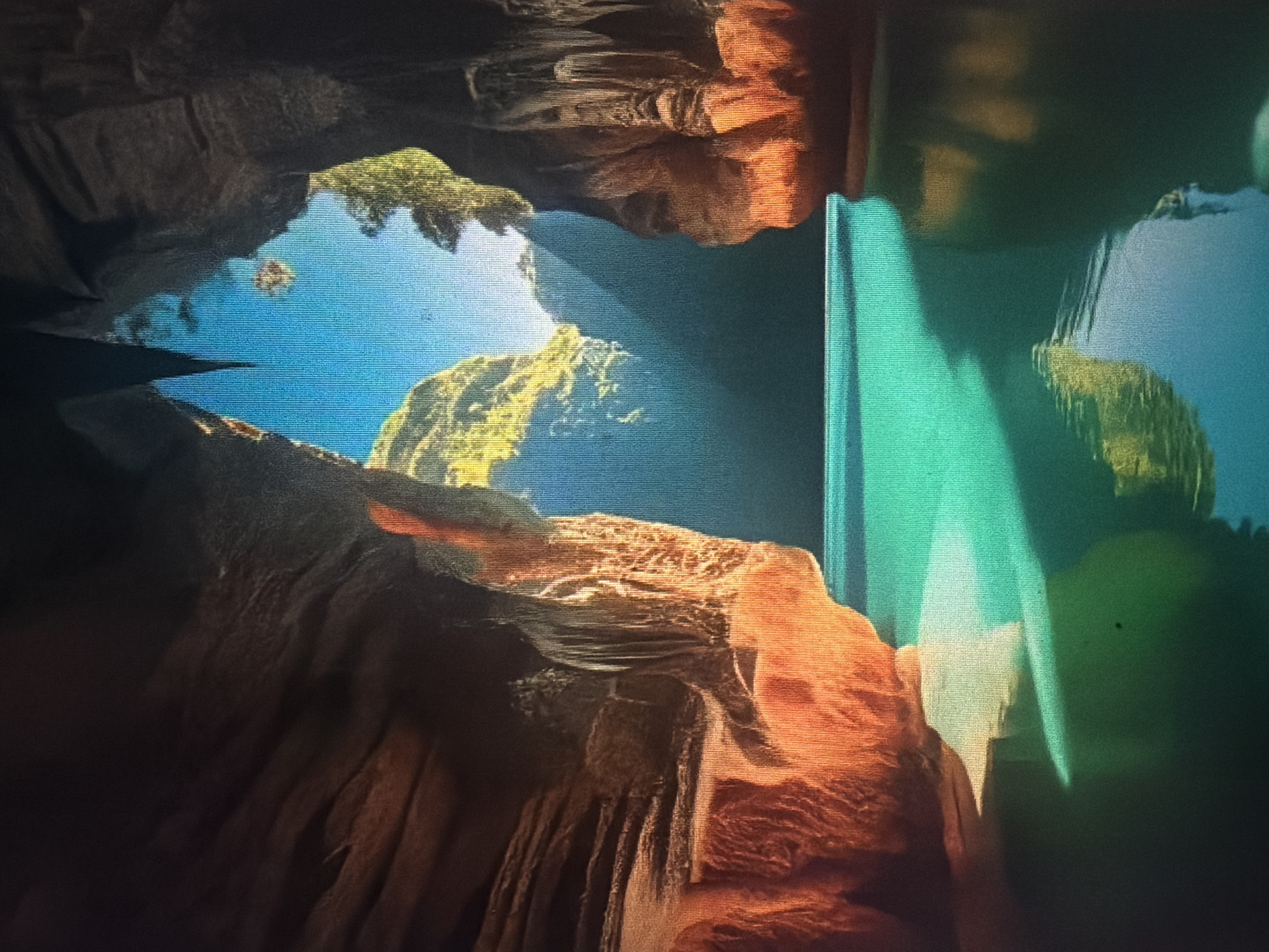 The image depicts the interior of a cave opening to a bright, tropical landscape. The cave walls are rugged and textured, with dramatic rock formations in shades of orange and brown. The cave opens into a body of turquoise water, which is calm and reflects the light. In the distance, sunbeams pierce through a gap between cliffs, illuminating lush green foliage on steep, rocky inclines. The sky is a vibrant blue. There are no people or man-made objects visible. The scene is evocative of a natural wonder, possibly a sea cave or a lagoon accessible through a cave. While the description mentions "Baan Mae Nam, Thailand," the image itself does not contain any specific visual cues that would definitively place it in that location.