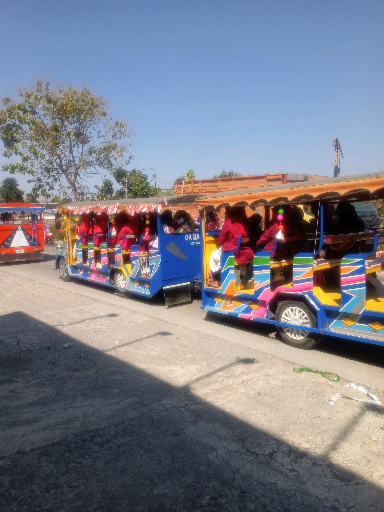 In Sumberlawang, Indonesia, two brightly colored, open-air jitneys, also known as "olinek" or "angkot" in Indonesia, are visible on a paved road under a clear blue sky. The jitney in the foreground is predominantly blue with vibrant yellow, pink, green, and white geometric patterns adorning its sides. It is carrying several passengers, most of whom are wearing red tops and headscarves. The jitney in the background is primarily orange and red with a white triangular emblem on its rear. It also appears to be carrying passengers. A large tree with sparse green foliage is visible behind the jitneys, along with some other greenery and structures that suggest an urban or semi-urban environment. The road surface is cracked in places, and a shadow is cast across it, indicating the presence of sunlight. Scattered on the ground near the foreground jitney are a few pieces of debris, including a green string-like object and some white plastic.