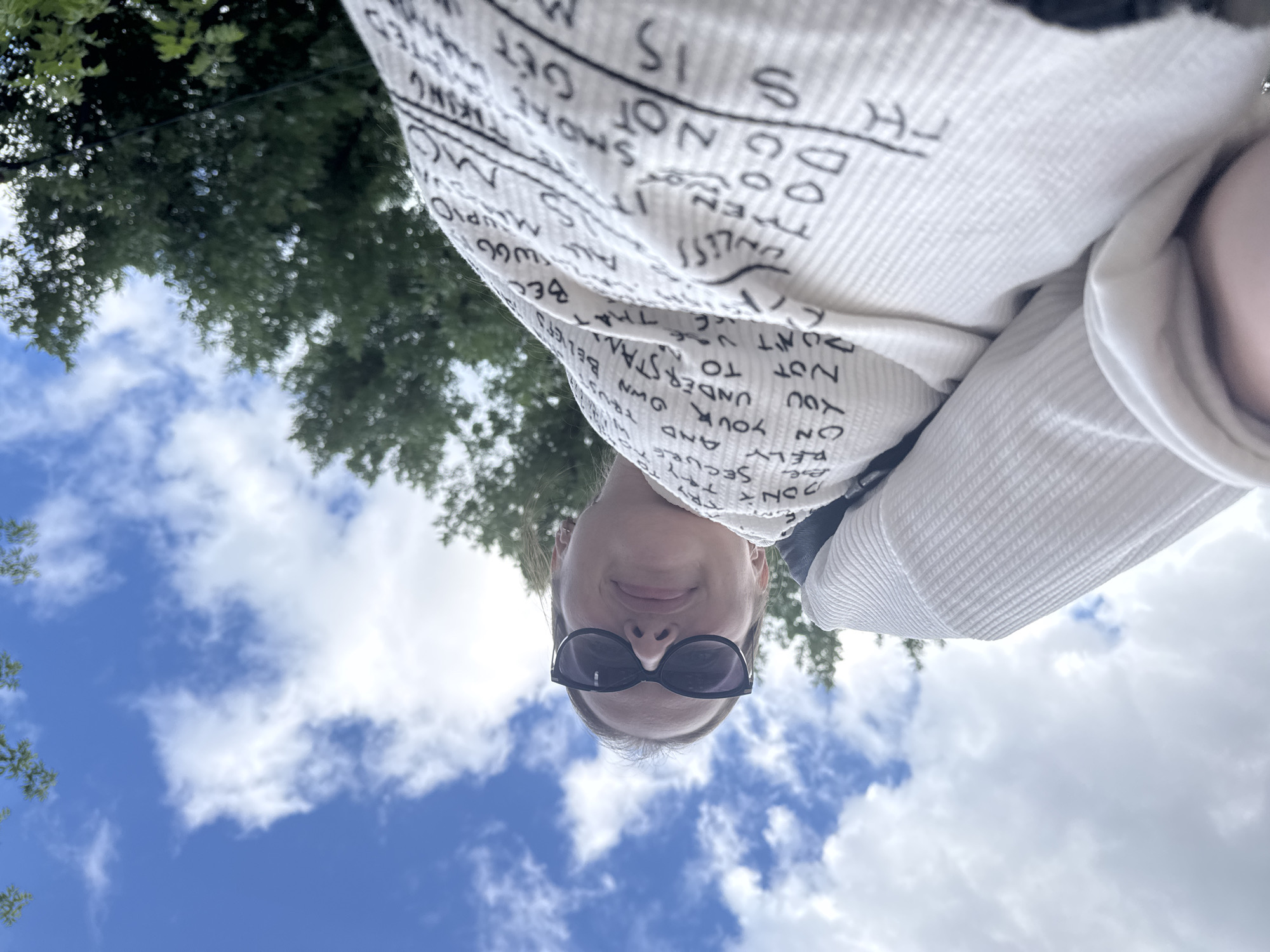 This is a first-person perspective, upside-down selfie taken outdoors, likely on a sunny day with fluffy white clouds in a bright blue sky. The subject is a person wearing sunglasses and a white top with text printed on it. In the background, there are green trees, suggesting a natural or park-like setting. The text on the shirt appears to be a mix of English words and possibly other languages or symbols. Some legible phrases include "BE SECURE," "RELY ON YOUR OWN," "DON'T TRUST THAT," and "SMOKE." The person is smiling, conveying a cheerful and relaxed emotion. The image appears to be taken during daylight hours, given the brightness of the sky. The location context suggests this might be Chaikovsky, Russia.
