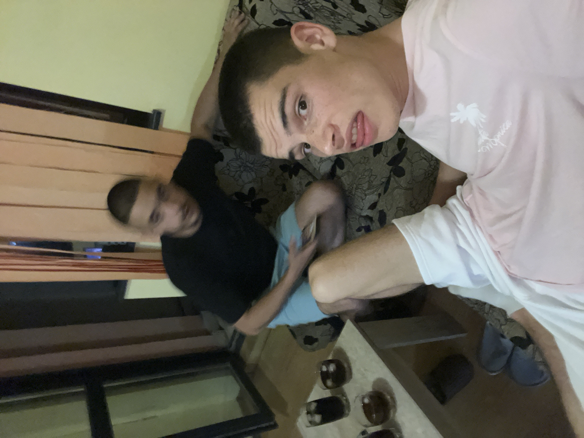 Two young men are sitting on a floral-patterned couch in an indoor setting. One is wearing a pink T-shirt with a graphic and white shorts, while the other is wearing a black T-shirt and light blue shorts. A table with several small glasses is visible near the bottom of the frame. A pair of blue shoes are on the floor next to the couch. Light filtering through a window, possibly a curtain, is visible on the left. The location is identified as Plovdiv, Bulgaria, although there is no visual confirmation of this in the image.
