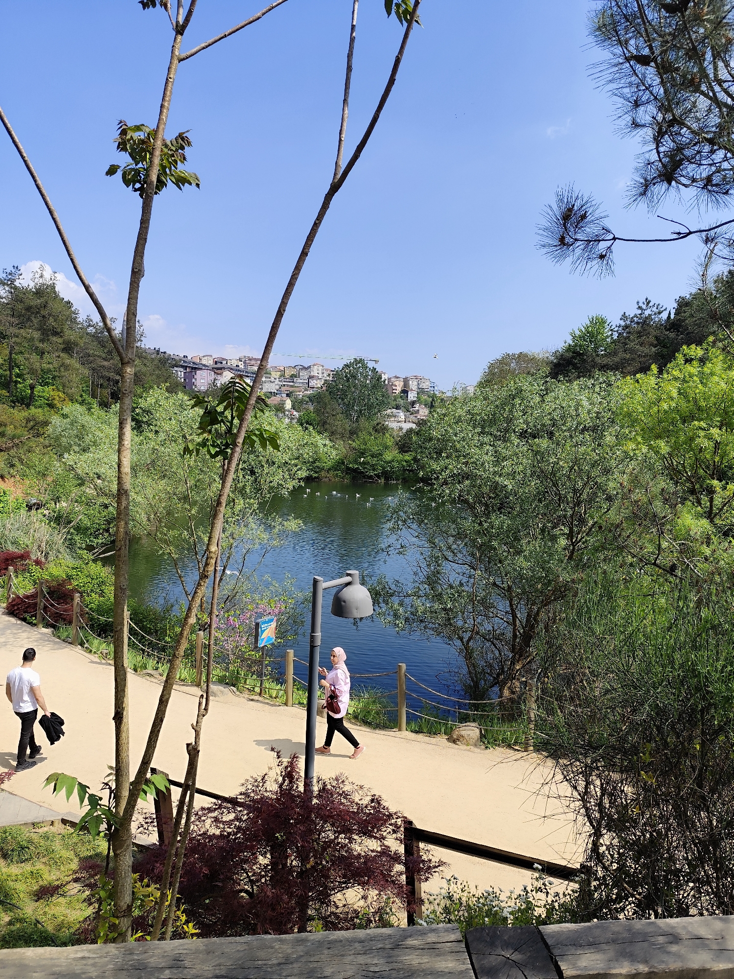 The image depicts a scene in a park located in Sarıyer, Turkey. A calm body of water, possibly a lake, is visible in the middle ground, reflecting the clear blue sky. Several buildings can be seen in the far distance.
A paved pathway runs along the side of the lake. A man in a white shirt and dark pants walks along the path, carrying a black jacket. A woman wearing a light pink headscarf and a light pink top walks in the opposite direction, looking at her phone. A gray lamppost is positioned near the woman on the pathway.
The scene is lush with vegetation. Trees line the pathway, with some branches and trunks visible in the foreground. The park environment includes a wooden railing along the pathway near the water's edge.