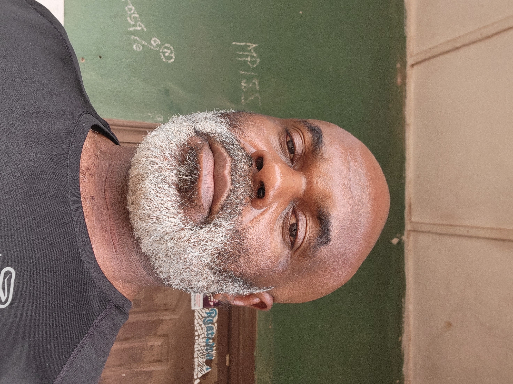 The image shows a medium-close shot of a Black man with a bald head and a gray beard. He is wearing a black sleeveless shirt. The background is a green wall with writing on it and a tan-colored wall with a seam. Below him, there is a dark brown doorway with a patterned piece of cloth visible. The location is potentially in Sabon Tasha, Nigeria. The subject's gaze is directed towards the camera. No actions or interactions are discernible in this static image.