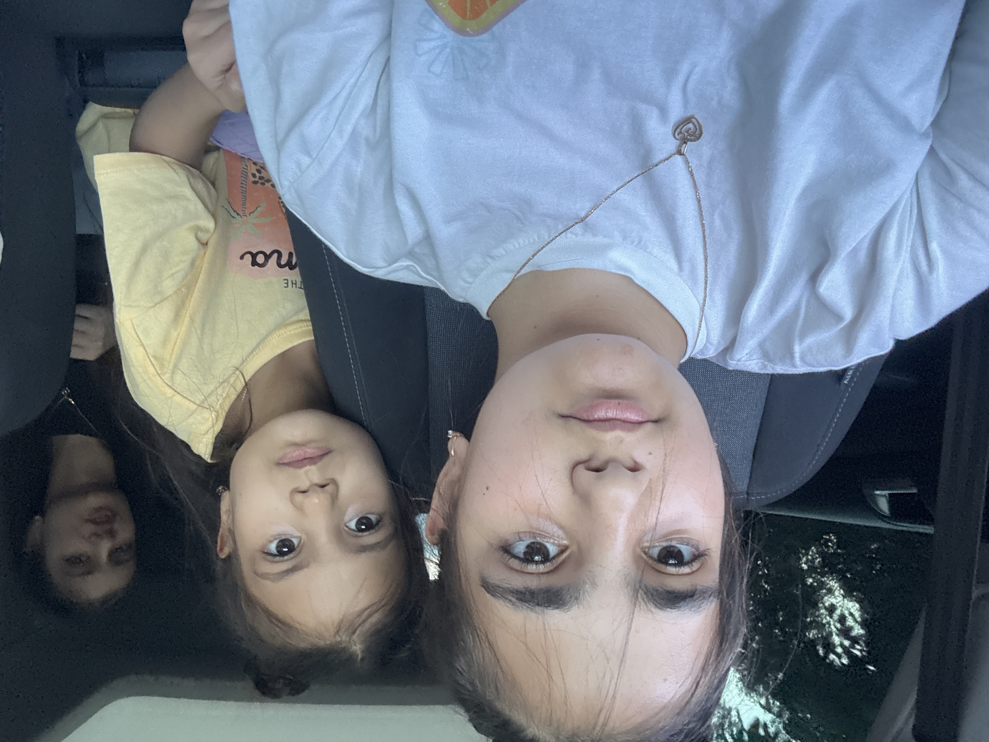 Three young females are inside a vehicle, likely a car, as suggested by the car seat and interior. The female in the foreground is looking directly at the camera, with her head and shoulders filling the frame. She has dark hair pulled back and is wearing a white t-shirt with a delicate silver necklace featuring a heart pendant. Behind her, slightly to the right, is another young female with dark hair styled in a bun, wearing a yellow t-shirt. She is also looking towards the camera. In the background, a third young female is visible, out of focus and to the far right, wearing a black t-shirt and a necklace. The interior of the car appears to be dark, with glimpses of dark upholstery and a dark headliner. Outside the car, through the windows, can be seen dark foliage, suggesting a wooded or tree-lined area. The lighting is somewhat dim, possibly indicating late afternoon or overcast conditions.
