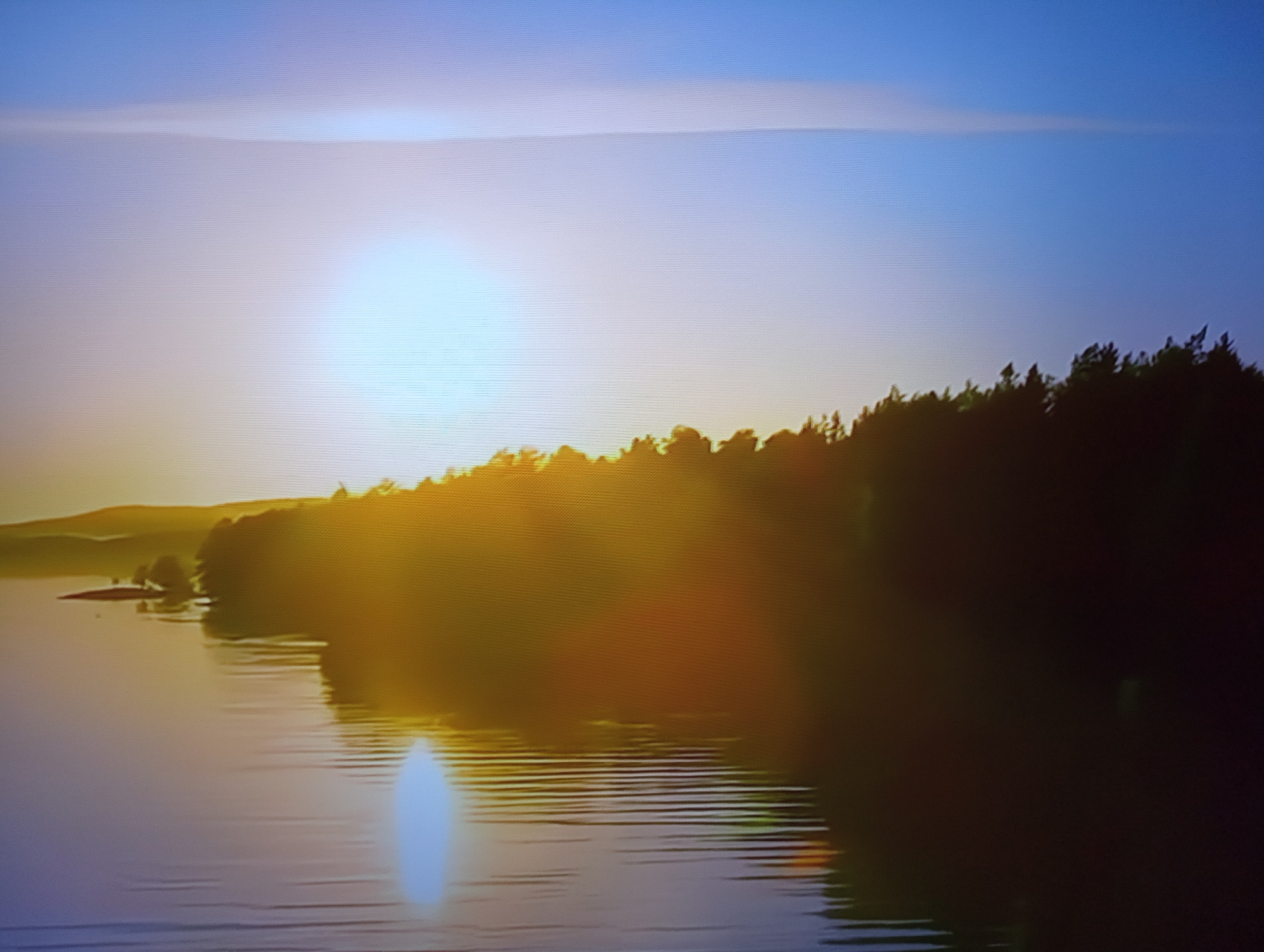 The image depicts a tranquil sunset over a body of water, likely a lake or river, with a tree-lined shore. The sun, a bright, hazy orb, is low on the horizon, casting a warm, golden glow across the scene. Its reflection shimmers on the rippling surface of the water, creating streaks of light. The sky transitions from a pale blue at the top to a softer, lighter hue near the horizon, indicating the late hours of the day.
The shoreline is densely forested with dark green trees, silhouetted against the bright sky. In the distance, gentle hills are visible, also bathed in the warm sunset light. A small, treed island or peninsula juts out into the water on the left side of the frame. The overall impression is one of peace and natural beauty, with no signs of human activity or man-made objects apart from the implied presence of a viewpoint from which the photo was taken. The time of day is clearly sunset. The weather appears calm, with the water showing only gentle ripples. Given the location context of Dnipro, Ukraine, this scene could be a view along the Dnipro River or one of its reservoirs. There is no visible text in the image.
