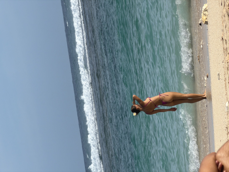 The image presents a beach scene in Ungasan, Indonesia, under a clear blue sky. A female subject, viewed from the back, stands at the edge of light-colored sand where it meets the turquoise ocean water. She wears a fuchsia-pink string bikini. Her dark hair is pulled back, featuring a white flower, possibly a frangipani, behind her right ear. Her left arm is raised, with her hand near her forehead, suggesting she is shielding her eyes while looking out towards the water. Small waves break at the shoreline, while a prominent line of larger white-capped waves is visible further out in the darker blue ocean, indicative of a reef or distant swell. The horizon line is observably tilted, rising from left to right. In the bottom-left foreground, the partial view of another person's tanned legs and arm is visible, suggesting they are seated or reclined on the sand.