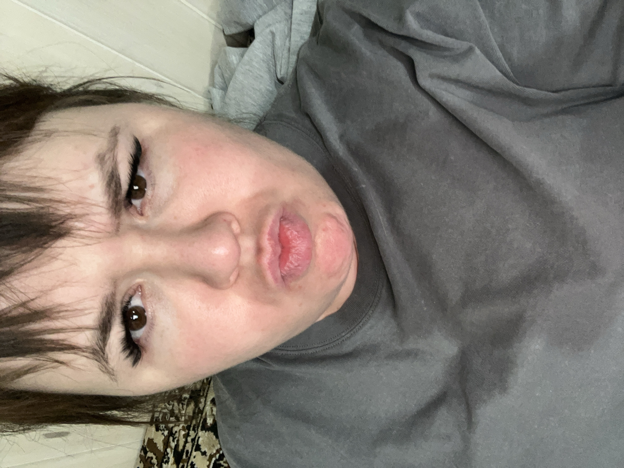 A close-up, tilted, selfie-style image shows a person with fair skin, brown eyes, and dark hair. The person is pouting their lips. The person appears to be wearing eyeliner and possibly false eyelashes. They are wearing a grey top, and a lighter grey garment is visible behind their head. The background includes a white, paneled wall and a patterned surface. There is no identifiable information to confirm the location is Chaikovsky, Russia.