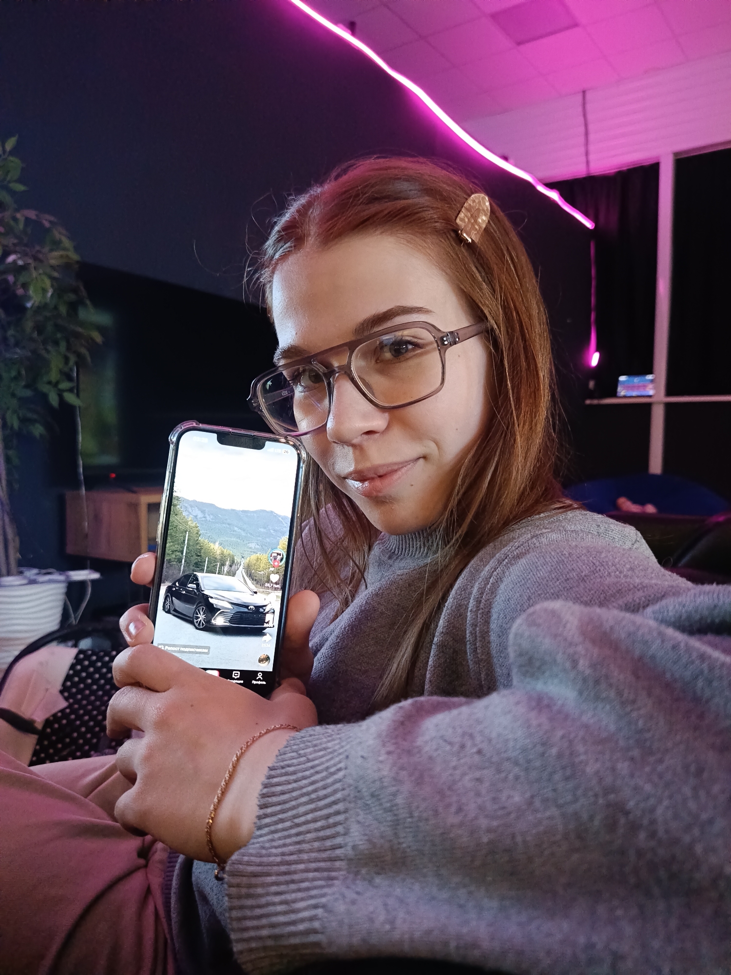 A young woman with shoulder-length reddish-brown hair and large, clear-framed glasses is featured in a close-up, casual indoor setting, possibly in Chaikovsky, Russia. She has a slight, friendly smile and looks directly at the camera. She wears a grey, ribbed long-sleeved sweater and light pink trousers. A small, golden, textured hair clip adorns the right side of her head, and a thin golden chain bracelet is on her left wrist.
She holds a clear-cased smartphone in her left hand, angling it to display its screen. The phone shows a video or image of a black sedan (resembling a Toyota Camry) parked on the side of a winding road, with mountains and green foliage in the background under a bright sky. The screen displays elements of a social media interface, including icons for likes, comments, shares, and a profile. Visible Cyrillic text at the bottom left reads "2. Pост подписчиков" (2. Subscriber growth), and "23,3К" (23.3K) is also visible.
The background is dimly lit, dramatically accented by bright pink neon strip lights tracing lines across the ceiling and down a wall, creating a modern and vibrant atmosphere. A dark curtain or wall is behind her, with hints of a plant in a pot and wooden furniture to the left. The scene suggests an evening or nighttime setting due to the artificial lighting. The woman appears to be presenting the content on her phone while being photographed.
