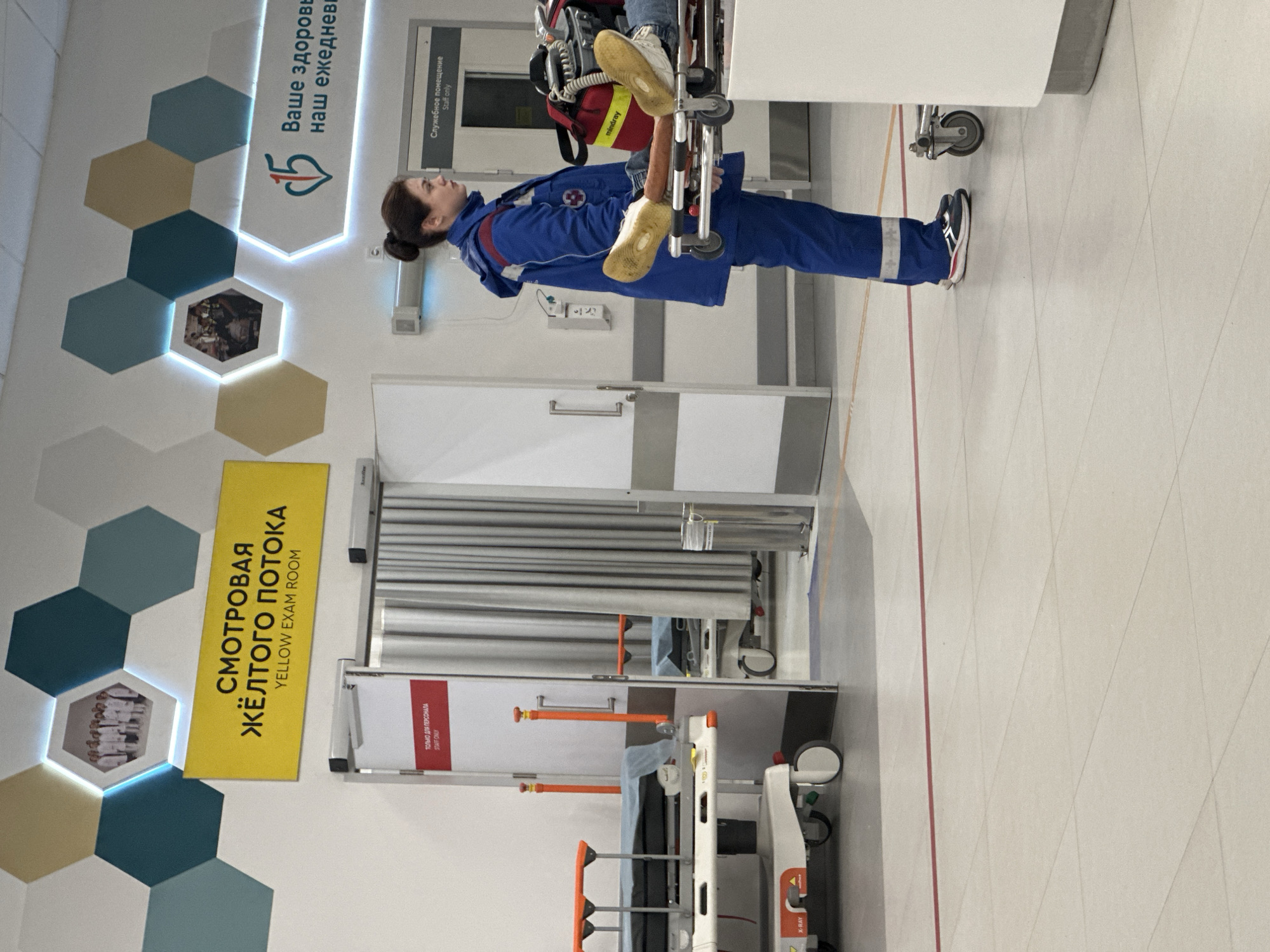 The image shows the interior of a medical facility. A female paramedic in a blue uniform is standing next to a stretcher. A sign on the wall reads "YELLOW EXAM ROOM". Another sign has Russian text and the number "15." There are hexagonal designs on the wall. A staff-only sign is visible. Text on medical equipment says "Mindray." The location is indicated by the Russian language signs and architectural style, which suggest it is Moscow, Russia.