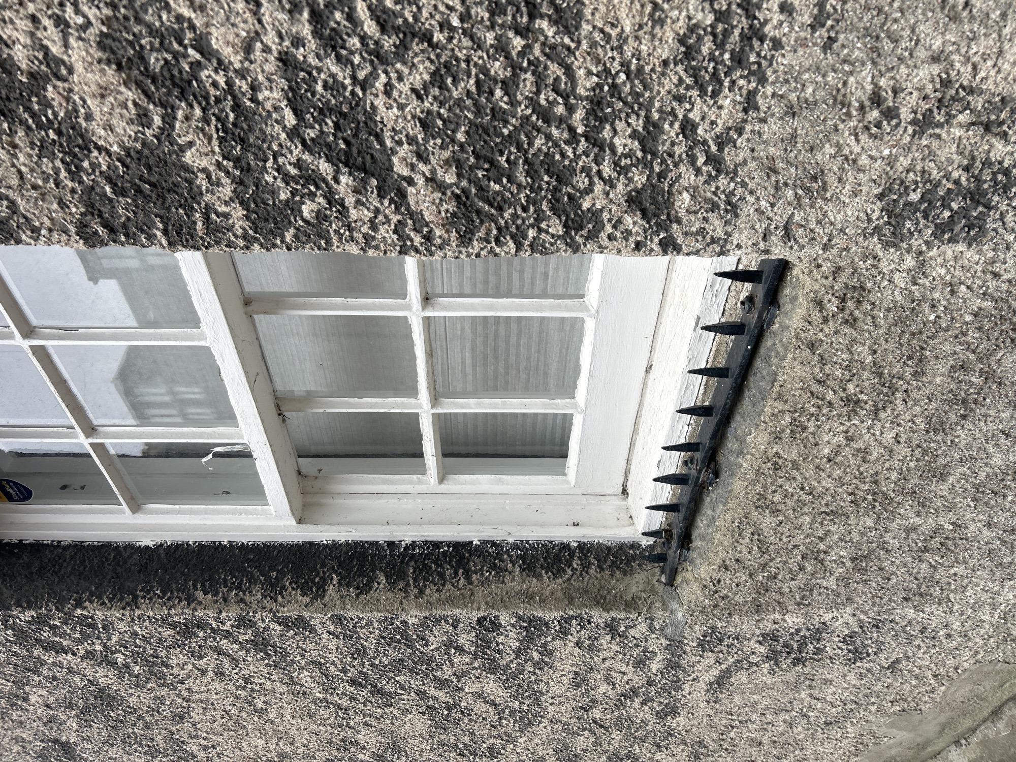 Captured in Aberdeen City, United Kingdom, the image presents a close-up view of a window set within a textured building facade. The window, painted white, features a grid pattern of panes and appears slightly aged, with minor signs of wear such as small cracks. To the right of the window, a black metal deterrent device with protruding spikes is affixed to the building's exterior, presumably to discourage birds or other animals. The building's facade exhibits a rough, speckled texture in varying shades of gray, contributing to an overall impression of age and weathering. The composition suggests a static scene focused on architectural details.