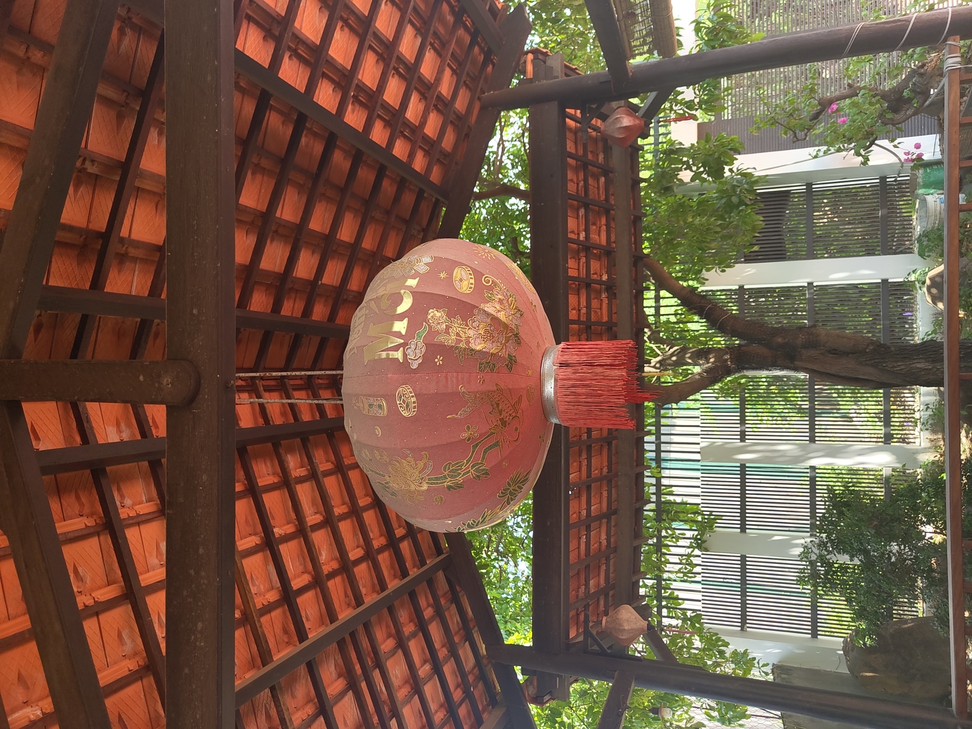 A red lantern hangs from the wooden beams of a tiled roof structure. The lantern has gold Chinese characters and floral designs. A fringe of red tassels hangs from the bottom. The structure is outdoors, with trees and a fence visible in the background. The scene is set in Bac Nha Trang Ward, Vietnam.