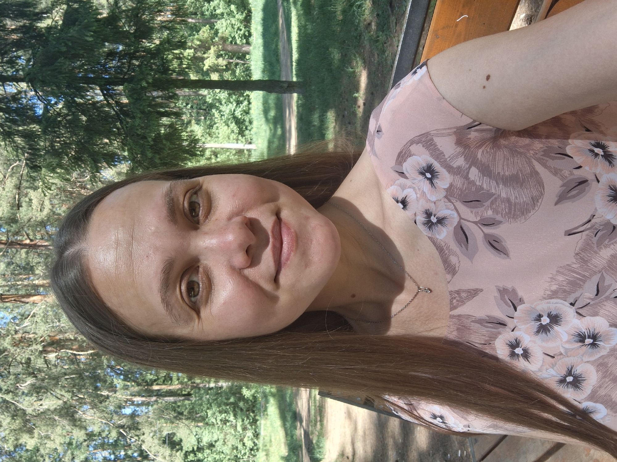 A woman with brown hair is smiling at the camera. She is wearing a light pink floral shirt and a silver necklace. The background shows a park with green trees and a path. The location is identified as the city of Chaikovsky, Russia. The woman is sitting on a wooden bench.