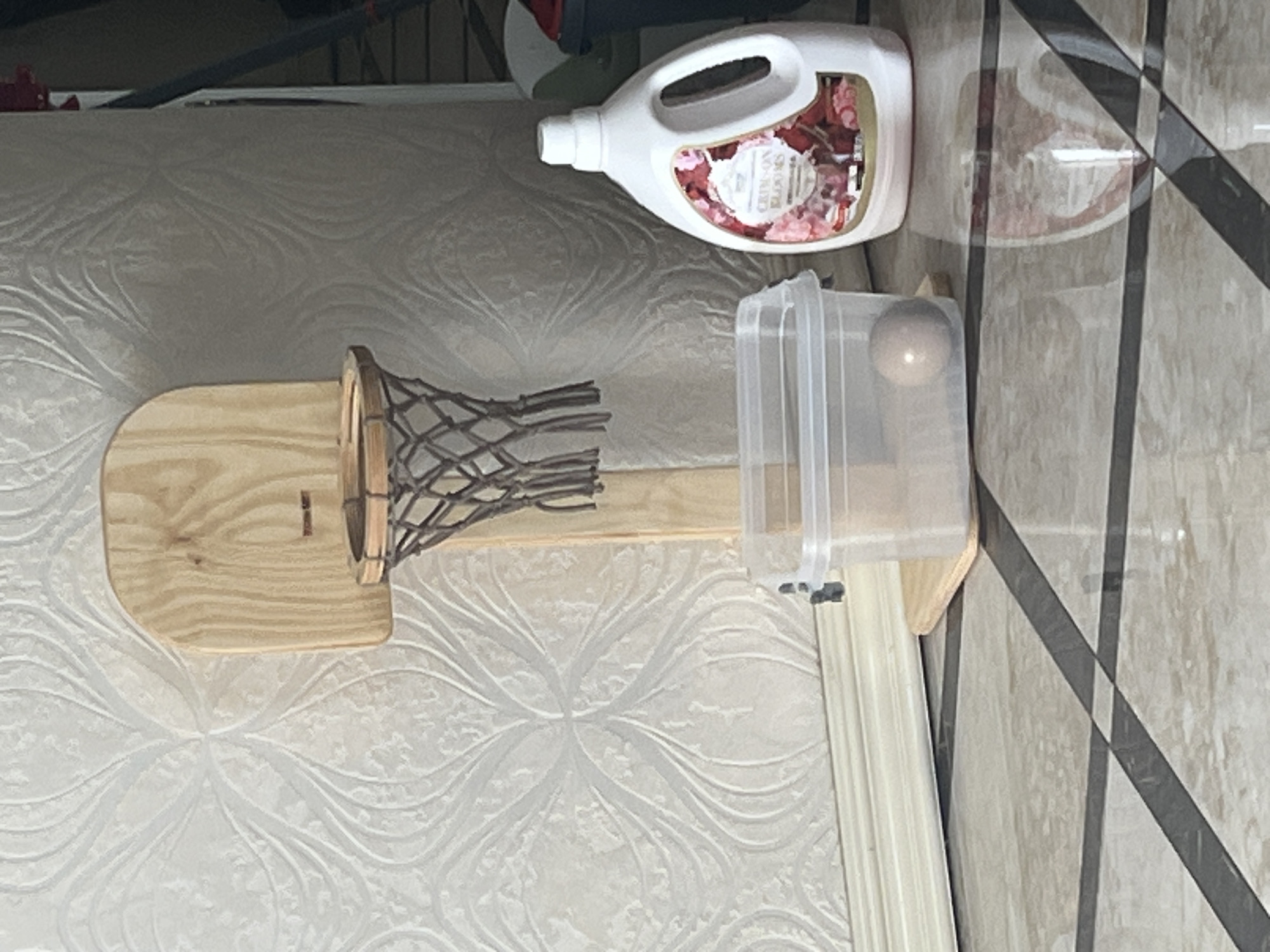 An indoor scene depicts a small wooden basketball hoop with a net, positioned against a textured, light-colored wallpaper. To the right of the hoop, a large white bottle of laundry detergent with a floral label stands on a tiled floor. In front of the detergent bottle and hoop, a clear plastic container holds a single light-colored ball. The floor is tiled with a pattern of alternating light and dark rectangular tiles. The scene appears to be a domestic setting, possibly a room in an apartment or house. There are no visible people. The overall impression is one of a staged or intentionally arranged display of household items.