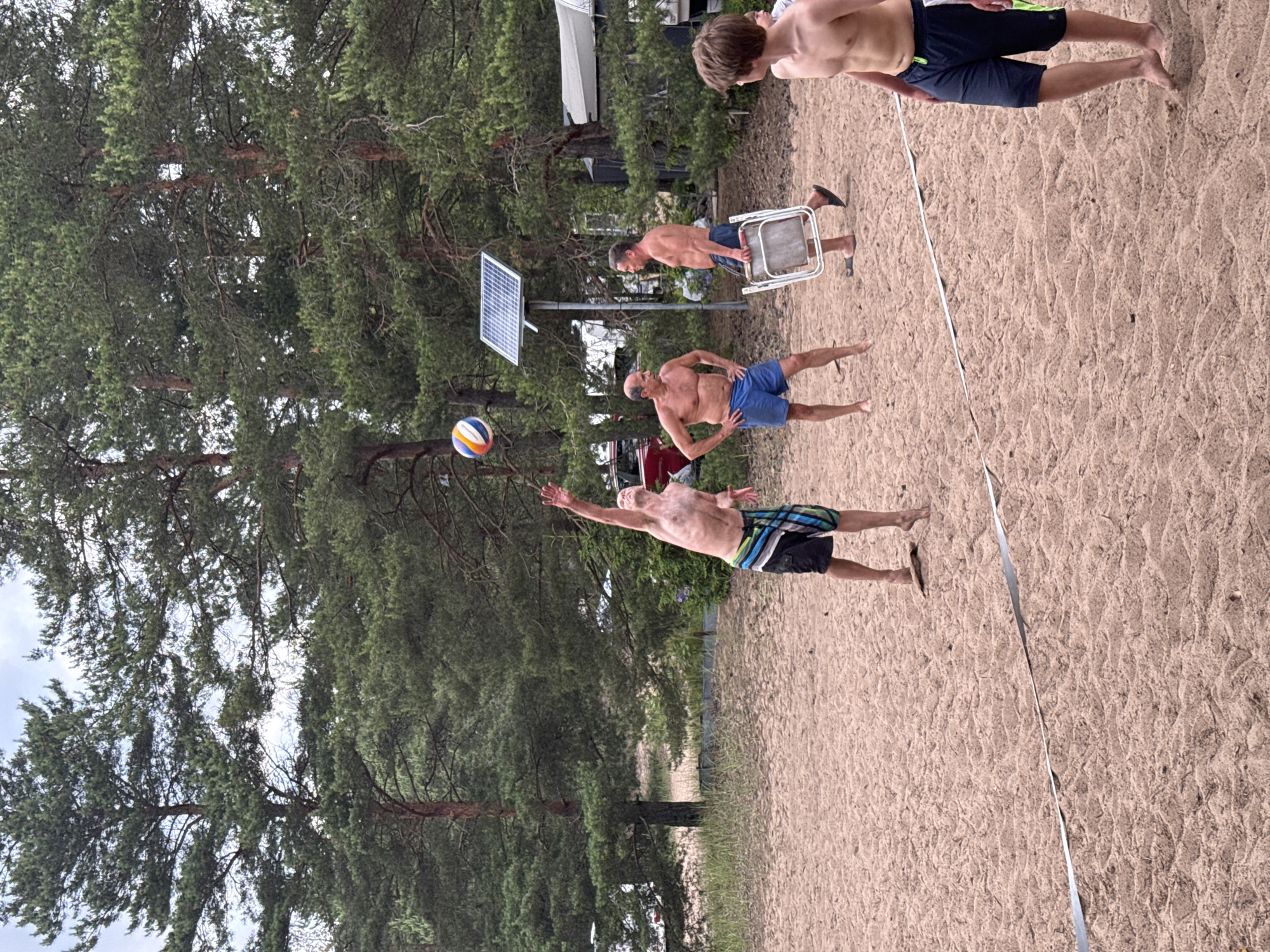In Zelenaya Roshcha, Russia, on a sandy beach with tall pine trees in the background, four individuals are playing volleyball. A man in checkered shorts is mid-jump, reaching for the ball, which is visible in the air. To his right, another man in blue shorts stands watching, while a third man, also shirtless, carries a folded chair. In the foreground, a young person in dark shorts stands facing away from the camera, towards the volleyball game. A white line demarcates the court on the sand, and a solar panel is mounted on a pole behind the players. The scene suggests a casual outdoor recreational activity.