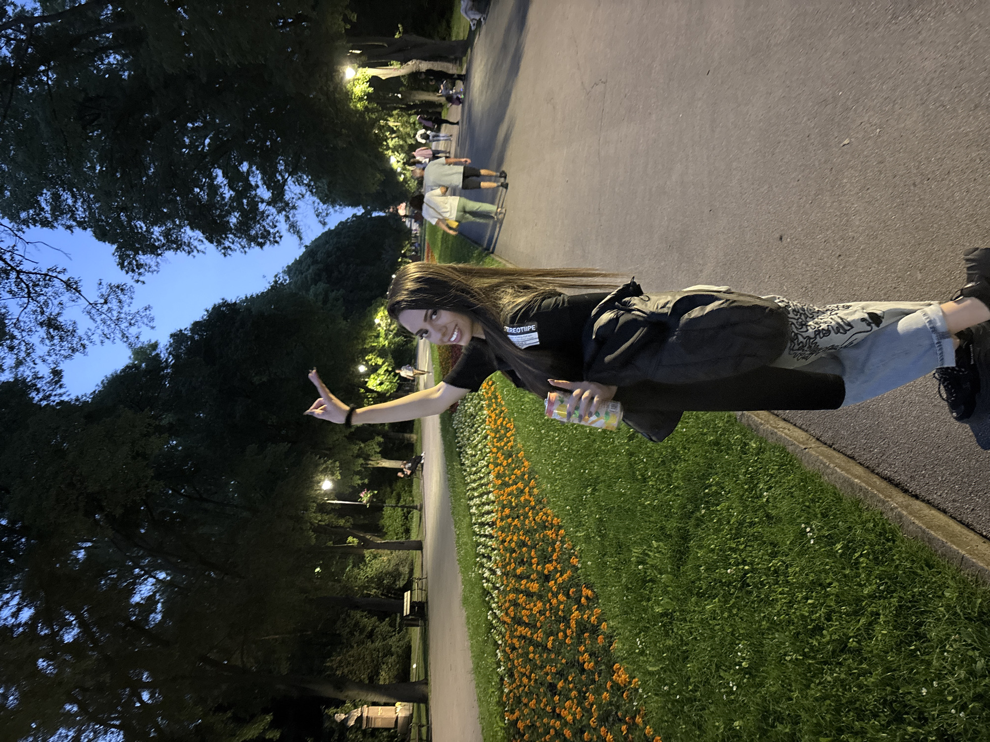 The image captures a young woman posing in a park or public garden in Varna, Bulgaria, during the evening. She stands on the edge of a paved pathway, with a vibrant flowerbed of orange and white flowers alongside green grass to her right.
The main subject, a young woman with long, dark hair, smiles at the camera, making a "peace sign" gesture with her left hand. Her right hand holds a colorful drink can. She is dressed in a black t-shirt, on which the word "STEREOTYPE" is clearly visible, light-colored jeans with a subtle graphic pattern, and dark shoes.
In the background, a paved path stretches into the distance, lined by tall, dark trees and illuminated by several streetlights. Other individuals are visible walking or standing further along the path. The sky is a deep blue, indicative of dusk or early night, providing a soft backdrop to the scene. The overall atmosphere appears relaxed and pleasant, set in a well-maintained public space.