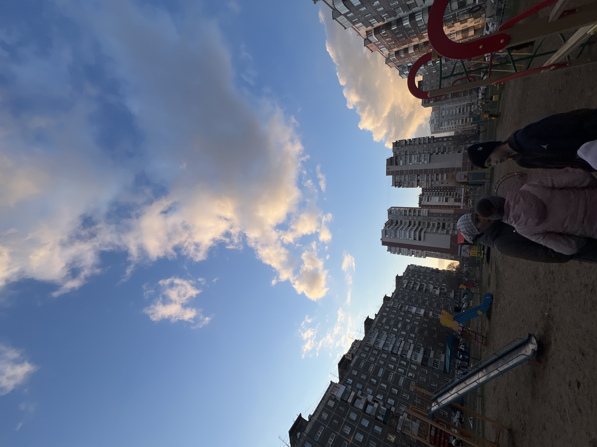 The image, oriented horizontally, depicts an urban playground scene in Perm, Russia, under a partly cloudy sky during late afternoon or early evening. The low sun casts a warm, golden light on the edges of some clouds and the facades of distant buildings, while other parts of the sky remain a clear blue.
In the foreground and to the right, two individuals are partially visible. One appears to be an adult or older child, dressed in a dark jacket and a black beanie. Next to them is a younger child in a pink puffer jacket and a light-colored beanie. Their faces are not clearly visible, and they seem to be looking down or away from the camera, present in the playground setting.
The playground area, covered in sand or dirt, features several pieces of equipment. A prominent red metal structure, possibly part of a climbing frame or swing set, is visible in the mid-ground. Further back, a blue and yellow plastic slide and a longer, older-looking blue metal slide can be seen.
The scene is framed by multiple tall residential apartment buildings in the background and along the left and right edges. These multi-story structures, with their numerous windows and balconies, appear to be typical urban blocks, contributing to the enclosed, city environment. The overall impression is one of a typical residential area with recreational facilities. No discernible text is visible in the image.