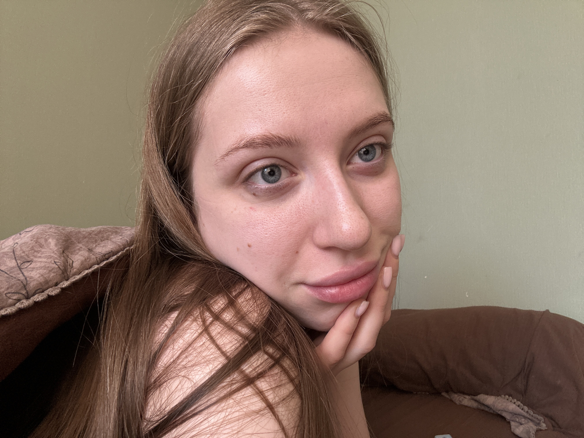 A young adult female, with fair skin, light blue eyes, and long, straight light brown hair, is positioned in a close-up, eye-level indoor portrait. She is reclining or lying down, her head tilted slightly, and her right hand is cupping her chin/jawline. Her fingernails are manicured with a light pink polish. A small mole is visible on her upper right cheek, near the corner of her mouth, and another on her forehead. She is looking directly at the viewer with a neutral to slightly smiling expression. Her left shoulder and upper torso are partially obscured by a textured brown pillow or blanket featuring a faint dark floral pattern. To the right of the frame, a plain brown upholstered surface, possibly a sofa or bed, is visible. The background consists of a plain, light olive-green wall. The specific location within Chaikovsky, Russia, cannot be determined from the visual information provided in the image.