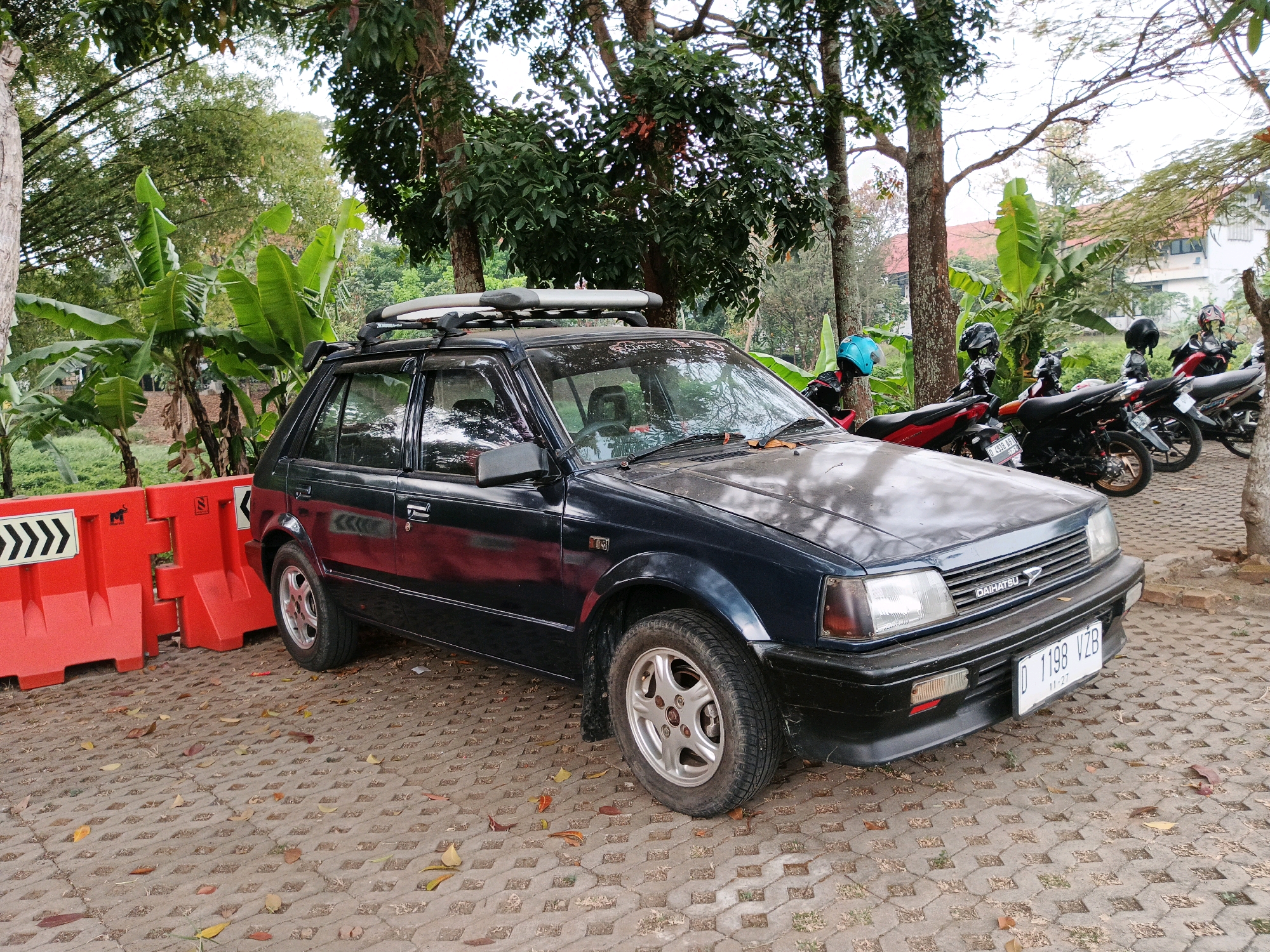 A dark blue, older model hatchback car is parked on a gray paver surface in what appears to be a parking lot or roadside area in Bandung City, Indonesia. The car has a roof rack with what looks like a surfboard or similar long, flat object on top. Behind the car, several motorcycles are parked, some with helmets on them. To the left of the car are bright orange traffic barriers. The environment is lush with green trees and vegetation, indicating a tropical or subtropical climate. The overall scene suggests a casual outdoor setting, possibly a public space or a private residence with a significant amount of greenery.