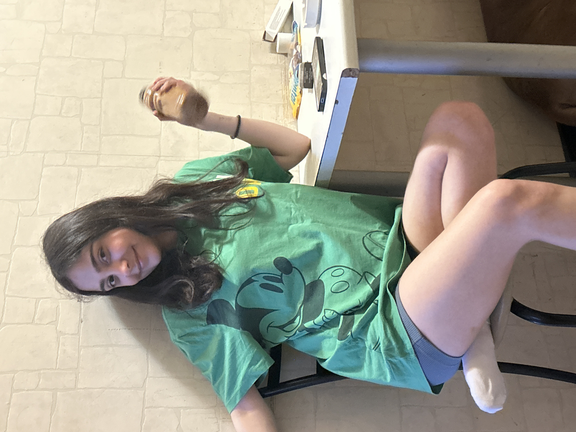 A young woman with dark hair is sitting on a black chair in front of a table. She is wearing a green t-shirt with a Mickey Mouse graphic and gray shorts. She is holding a glass jar in her right hand. The table has a smartphone and other items on it. The background is a cream-colored tiled wall.